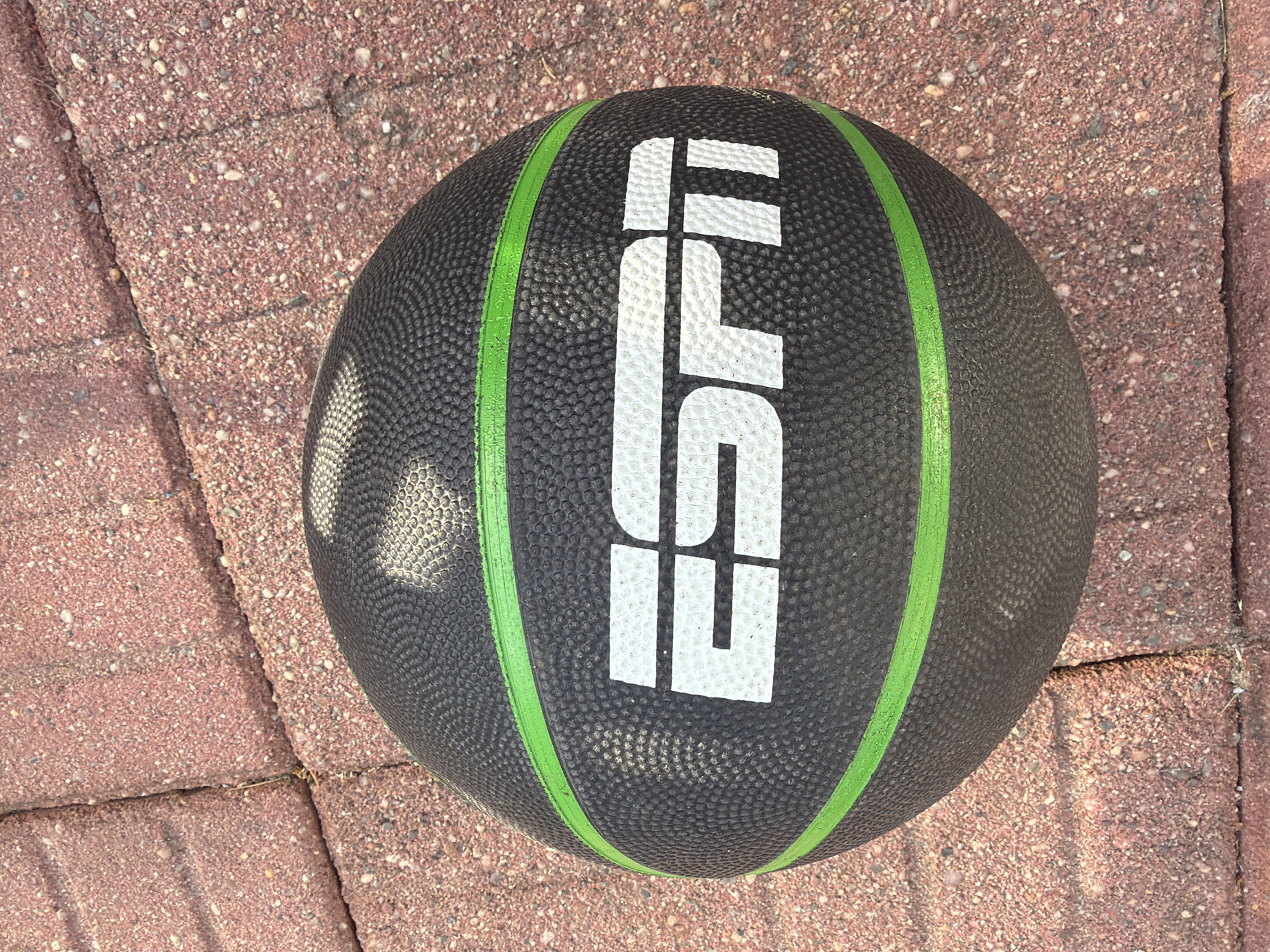 A black ESPN medicine ball rests on a red brick paver patio. The ball has two lime green bands running vertically around its circumference, and the word "ESPN" is printed in large white letters on its surface. The ball is positioned at an angle, with its lower left portion extending off the bottom of the frame and its upper right portion extending off the right edge. The bricks are a mix of red and brown hues, with visible grout lines and some small pebbles scattered on the surface. The lighting suggests it is daytime, with shadows cast by the ball and the surrounding environment. The scene appears to be an outdoor setting.