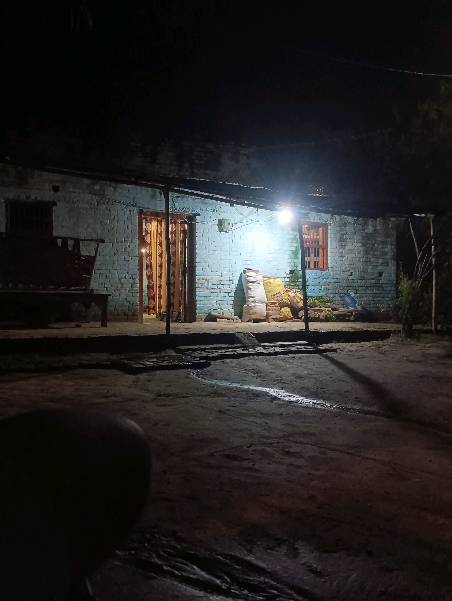 The image depicts an outdoor scene at night in Dakhin Parsa, India. A light illuminates the front of a blue brick house. In front of the house, there are several large sacks stacked, with what appears to be green produce or vegetation piled behind them. A window with wooden shutters is visible on the house. To the left, a wooden bench or seating area is situated in front of the house. The ground in the foreground is a dark, unpaved surface with some tire tracks and a wet patch reflecting the light. The overall atmosphere is dimly lit and rural.