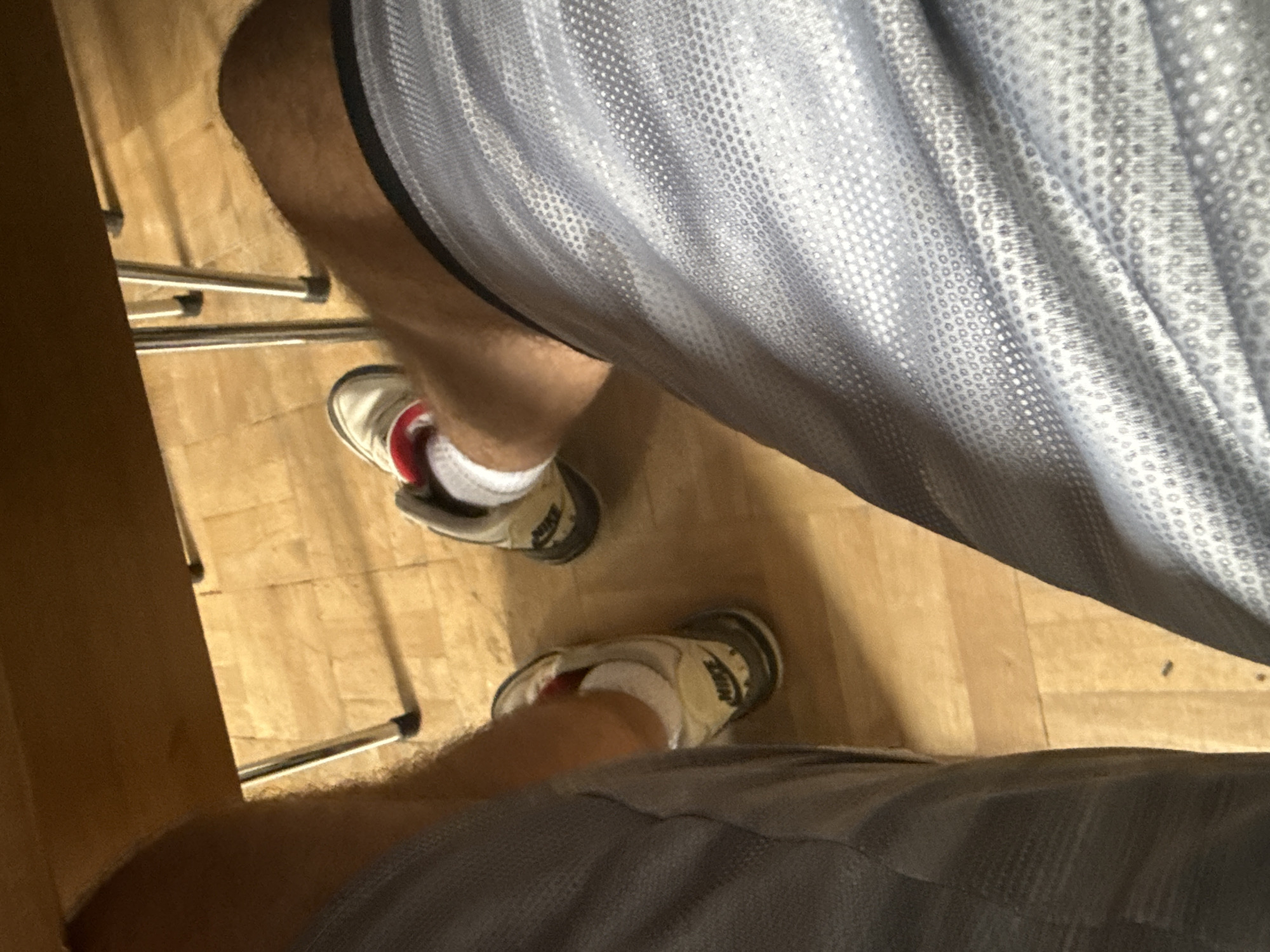 The image shows a close-up, low-angle view of a person's legs and feet, seated at a table. The person is wearing grey athletic shorts with a subtle patterned texture and white socks. Their feet are clad in Nike athletic shoes, one of which is white with red accents, and the other is a lighter beige with black and red accents. The shoes are resting on a light-colored wooden floor, which appears to be made of planks. In the background, the legs of a table or chair, made of shiny metal, are visible, along with the underside of a wooden table surface. The lighting suggests an indoor environment, likely during the daytime. There is no indication of the specific location in Sofia, Bulgaria, or any other identifying features of the city.