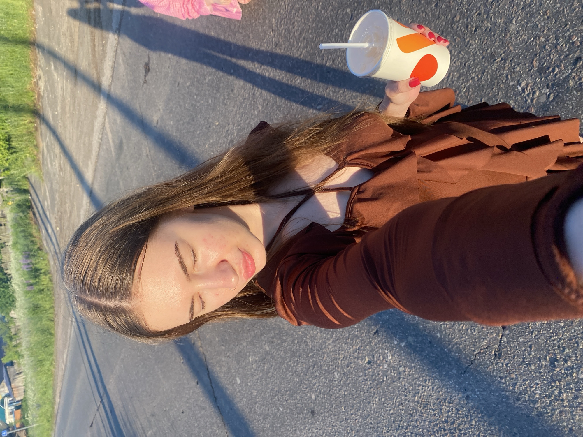 In Медведево, Russia, a young woman with long brown hair stands on a paved road, holding a white and orange beverage cup with a white straw. She is wearing a brown ruffled dress. Her eyes are closed, and she is looking down. Sunlight casts long shadows across the road. In the background, a grassy verge with scattered purple flowers lines the road, leading to wooden fences and houses.