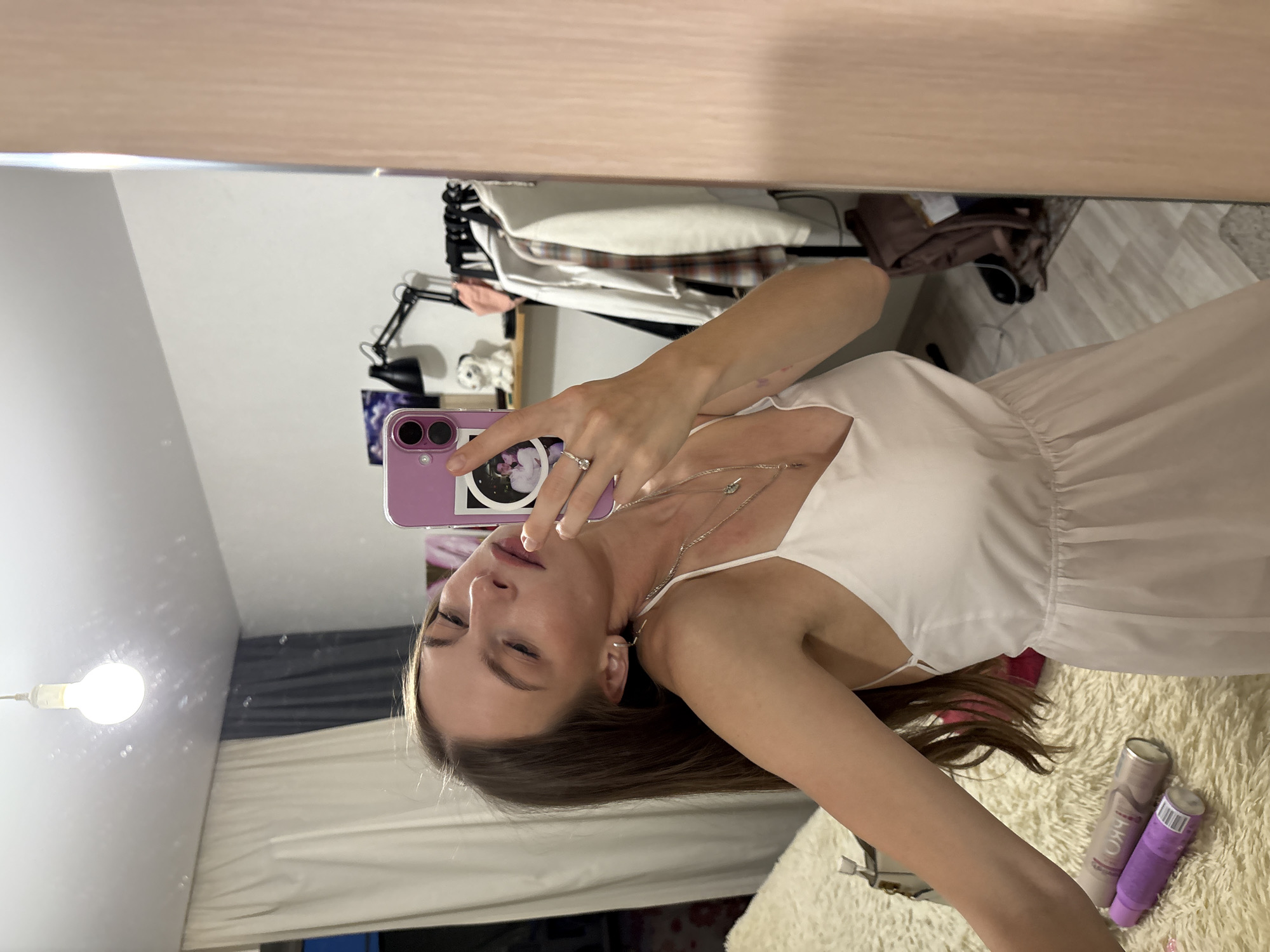 A young woman, seen from the chest up, takes a mirror selfie in what appears to be a bedroom or dressing area. She is wearing a light-colored, spaghetti-strap dress and has long, brown hair. She is wearing a ring on her right hand and a necklace. The background is a reflection in a mirror, showing a closet with clothing, a desk lamp, and a bare lightbulb. There are also two spray cans, one pink and one purple, visible on a fluffy rug in the lower right corner of the image. The overall lighting suggests it could be daytime or evening. There is no visible text in the image. The setting appears to be a personal space, likely a home.