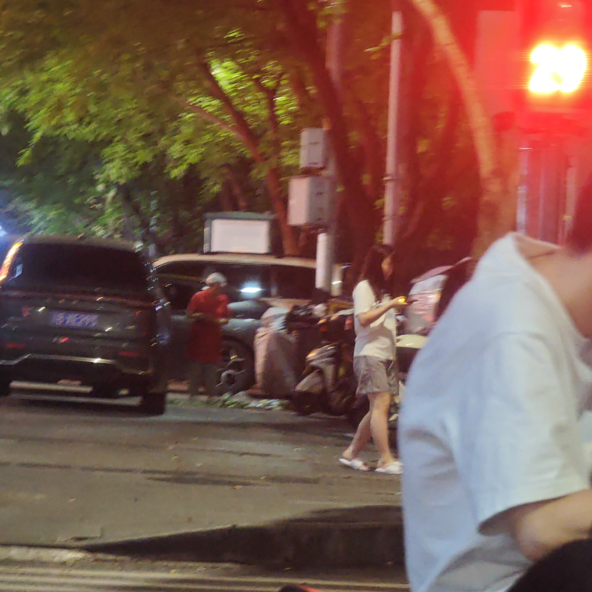 The image presents a street scene at night in what appears to be Yuzhong District, China. In the foreground, there is a blurry figure wearing a white shirt. In the middle ground, a woman with long hair is walking on the street, looking at her phone. She is wearing a white t-shirt and patterned shorts. Behind her is a parked scooter and a dark-colored SUV. Further back, a woman in red stands beside a light-colored car. Green trees line the street, illuminated by streetlights. A traffic light showing the number "28" is visible on the right side of the image. The street is paved, and there are white lines marking lanes. Overall, the scene suggests a typical evening in an urban environment.