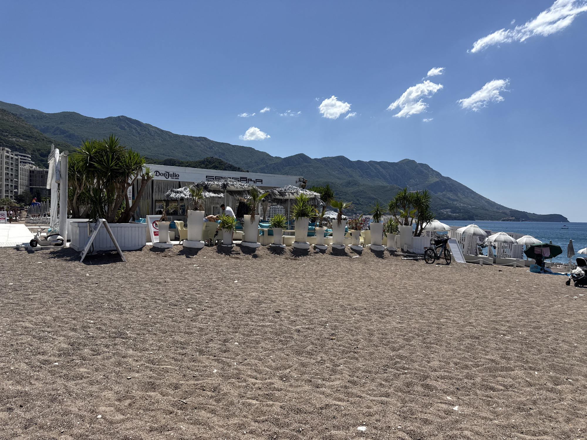 The image depicts a beach in Boreti, Montenegro, on a clear, sunny day. The foreground is dominated by a coarse, sandy beach with small pebbles. In the midground, a white beach bar or restaurant structure is visible, adorned with large white planters containing palm-like trees. The structure has a thatched roof and signage that includes "Don Julio" and "SPA & BAR". Several white umbrellas and beach chairs are set up in front of the bar. A white scooter is parked to the left of the bar, and a bicycle is leaning against a white structure on the right. A stroller is also visible on the far right. In the background, lush green mountains rise steeply, meeting a clear blue sky with scattered white clouds. The sea is visible to the right, meeting the shoreline. The overall scene conveys a tranquil beach atmosphere.