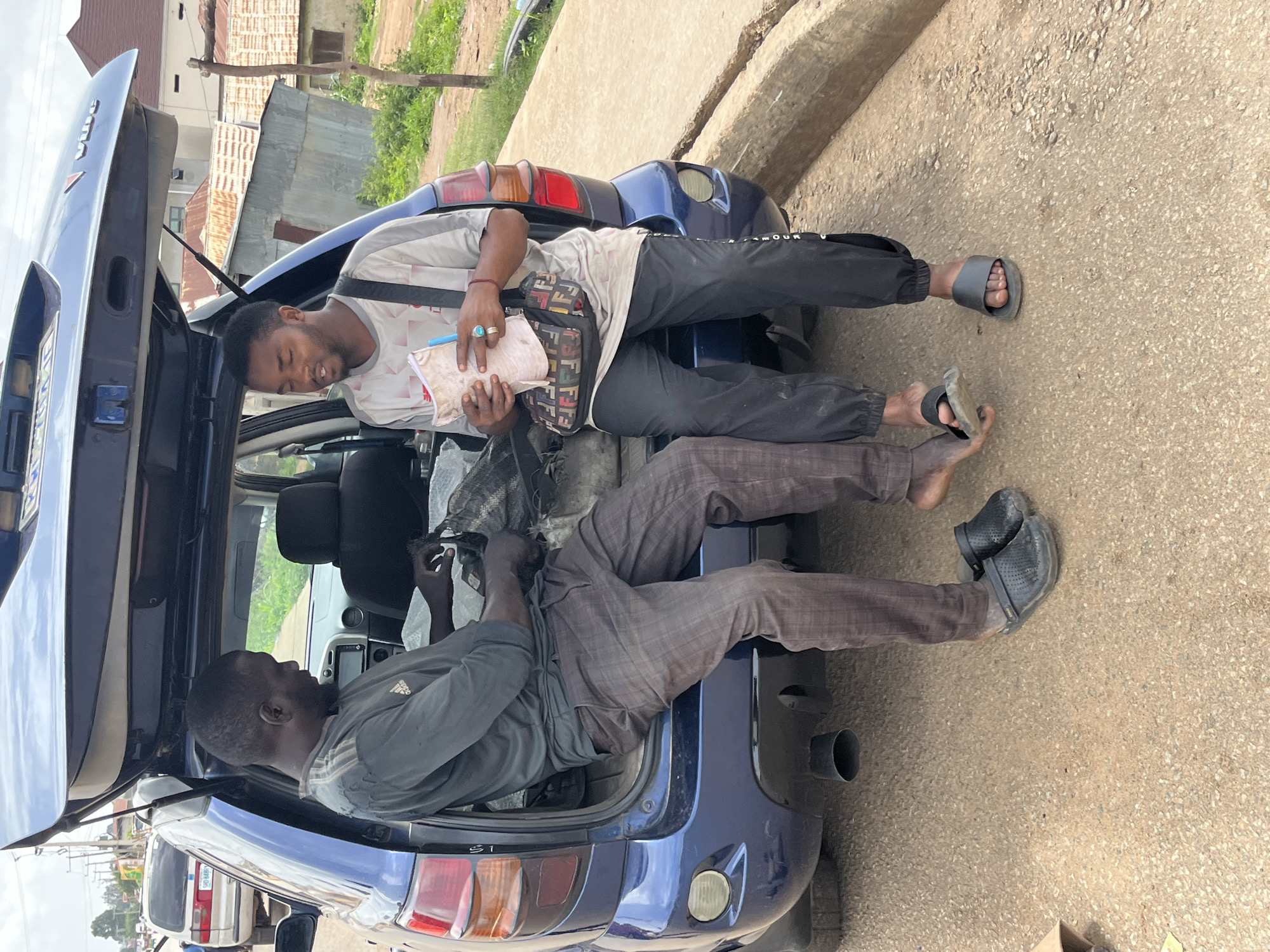 Two men are sitting on the rear bumper of a blue Toyota Matrix with its tailgate open. The man on the left, wearing a dark grey long-sleeved shirt and plaid trousers, is sorting through a black plastic bag with his hands. He is wearing black clogs. The man on the right, wearing a light grey t-shirt and black track pants, is holding a small notebook and a blue pen. He is wearing black slides and has a Fendi-patterned bag slung across his body. The car is parked on a dirt road next to a curb. In the background, there are buildings with corrugated metal roofs and some greenery. The scene appears to be in a residential or semi-urban area in Bukuru, Nigeria.