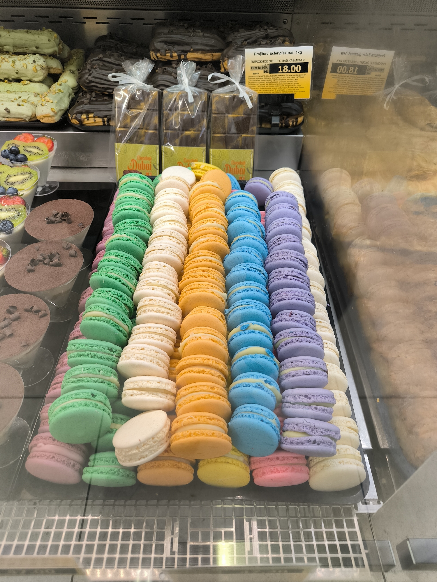 A retail display case filled with an assortment of pastries is shown in this close-up, eye-level shot. The display case appears to be part of a bakery or confectionary shop. In the foreground, rows of vibrantly colored macarons are neatly arranged in a vertical orientation. The macarons are stacked in columns, with colors including pink, green, white, yellow, blue, and purple. Behind the macarons, on the left side of the image, are several clear cups containing what appear to be desserts topped with fruit, such as strawberries, kiwi, and blueberries. To the right of the macarons, the display case continues with more pastries, including what look like eclairs covered in chocolate and pistachios. Further back on the right, a large pastry with a textured surface is visible, possibly a cake or bread loaf. Two yellow price tags are also visible, one in the middle left of the image with Russian text and a price of 18.00, and another on the right with Cyrillic text. The overall lighting is bright and artificial, typical of a retail environment, and the display case is made of glass. The background consists of the back wall of the display case, which appears to be a dark, metallic material. The scene is static, with no people or animals present. The specific location is not evident from the image itself.