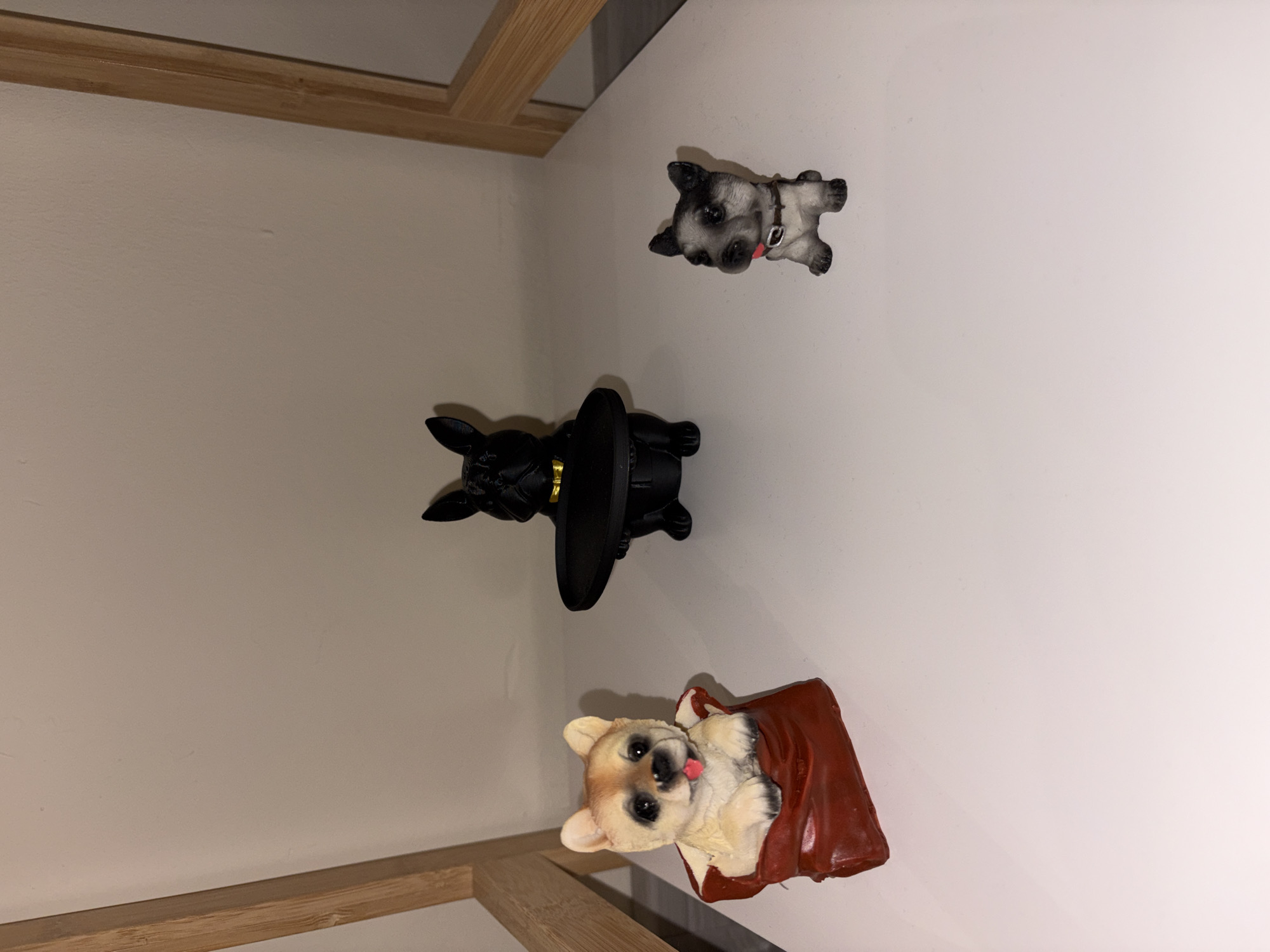 The image displays an indoor scene, located in Sofia, Bulgaria, featuring three decorative dog figurines positioned on a flat, white surface. Behind the figurines is a plain, light beige wall. Partial light-colored wooden frames are visible vertically on both the left and right edges of the image, suggesting a piece of furniture or shelving.
From left to right:
1. A light brown or beige dog figurine, resembling a puppy, is situated within a red, open, sack-like container. The figurine features black eyes and a small pink tongue slightly extended.
2. A black figurine, appearing to be a bulldog, stands in the center. It wears a metallic gold-colored collar and holds a black, circular, flat object, possibly a tray or dish, with its front paws.
3. A smaller gray and black dog figurine is seated on the right. It has a brown collar and a small red detail visible near its mouth, possibly a tag or tongue.
No people are present. The objects are static, indicating no active interactions or actions. The environment is clean and uniformly lit.