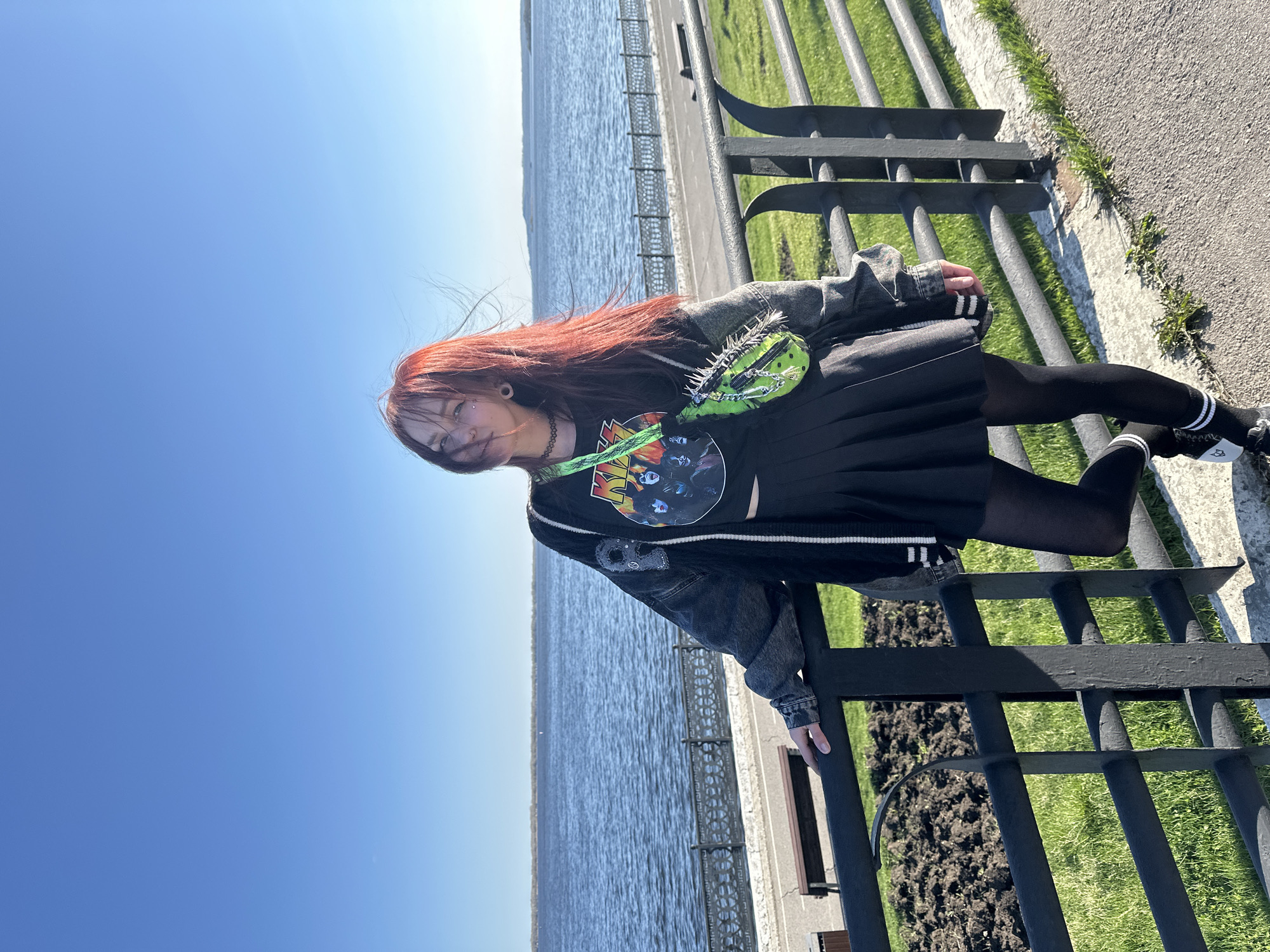 A young woman with long, reddish-brown hair stands outdoors on what appears to be a river embankment in Saratov, Russia. She faces slightly left, looking towards the camera with a neutral expression, and has ear gauges and a piercing above her lip. Her hair is gently wind-swept.
She wears a black t-shirt with a colorful graphic design, possibly a band or movie poster, underneath a dark grey or faded black denim jacket. A distinctive bright green fanny pack, heavily embellished with silver spikes and chains, is worn across her chest. Her attire is completed with a black pleated skirt, black tights, and black shoes paired with black socks featuring white stripes and small, cartoon-like character details. Her right hand rests on a black metal fence.
Behind the woman and the fence, a wide body of water, characteristic of the Volga River, extends horizontally under a clear, bright blue sky. A paved path runs along the waterfront. To the right of the woman, beyond the fence, there is green grass and patches of dark soil. The scene is brightly lit, indicating sunny weather during midday or early afternoon.