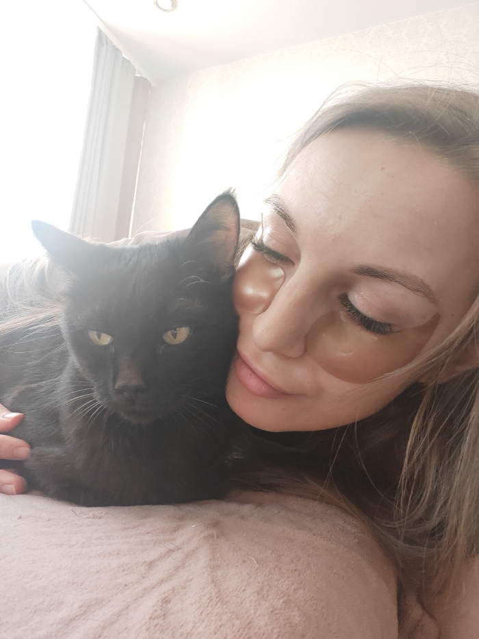 A blonde female and a black cat are depicted indoors in Chaikovsky, Russia. The female, positioned on the right, has closed eyes and two translucent, amber-colored patches applied beneath her lower eyelids. Her blonde hair is visible, and her left hand rests on the cat's back. The black cat, positioned on the left, has bright yellow-green eyes open and facing the viewer. Its head is immediately adjacent to the female's head. Both subjects are resting on a light-pink, textured fabric surface. The background is brightly illuminated, revealing a light-colored curtain on the left, a plain white wall, and a circular overhead light fixture partially visible at the top center. The scene is significantly backlit.