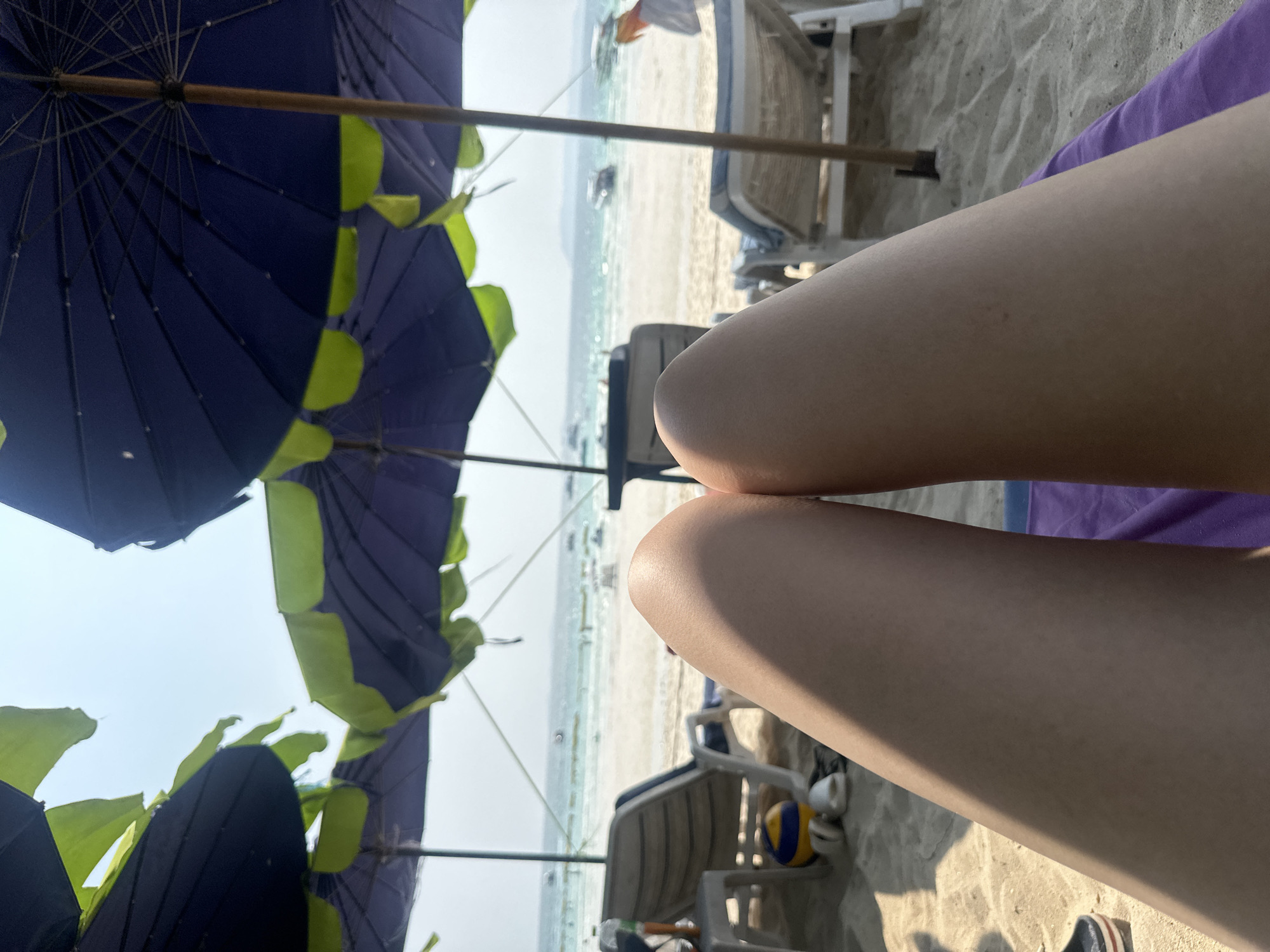 The image shows a beach scene in Pattaya City, Thailand. In the foreground are the legs of a person lying on a purple towel on a beach chair. Behind them are multiple beach umbrellas, blue and green, shielding the area from the sun. In the midground, the beach stretches toward the sea, with beach chairs and people scattered along the shore. Several boats are visible on the horizon in the background. The sky is pale, suggesting a sunny day. A volleyball is visible in the midground under a beach chair.