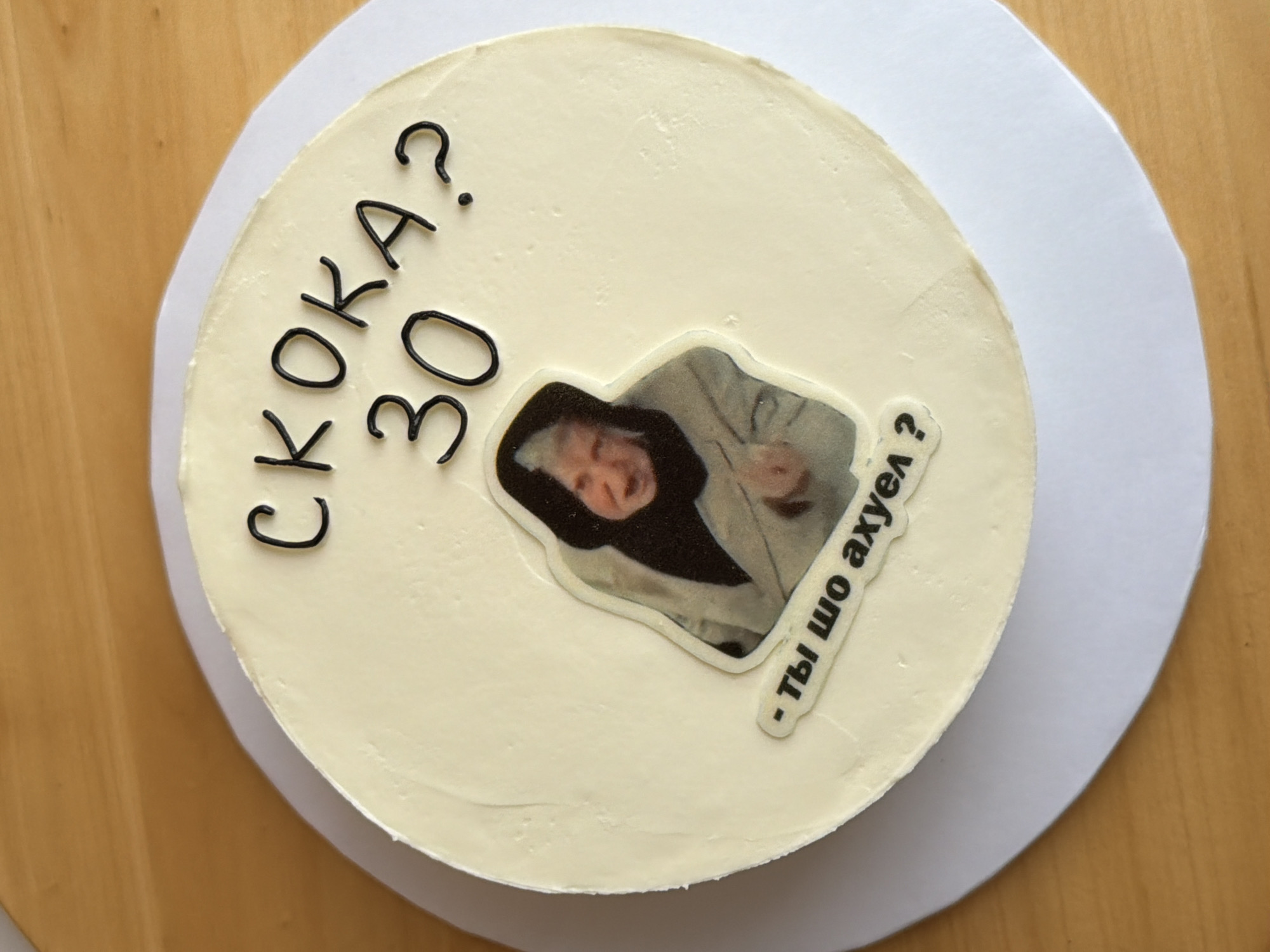 A top-down shot of a round cake on a larger white paper plate, placed on a wooden surface. The cake is covered in smooth, light-colored frosting. Written in black frosting are the words "СКОКА? 30?" to the left of center. A rectangular image featuring a person in a light-colored coat is adhered to the cake. To the right of the image is a smaller, rectangular sticker with the text "- ты шо ахуел?". There is no visible indication of Tel-Aviv, Israel.