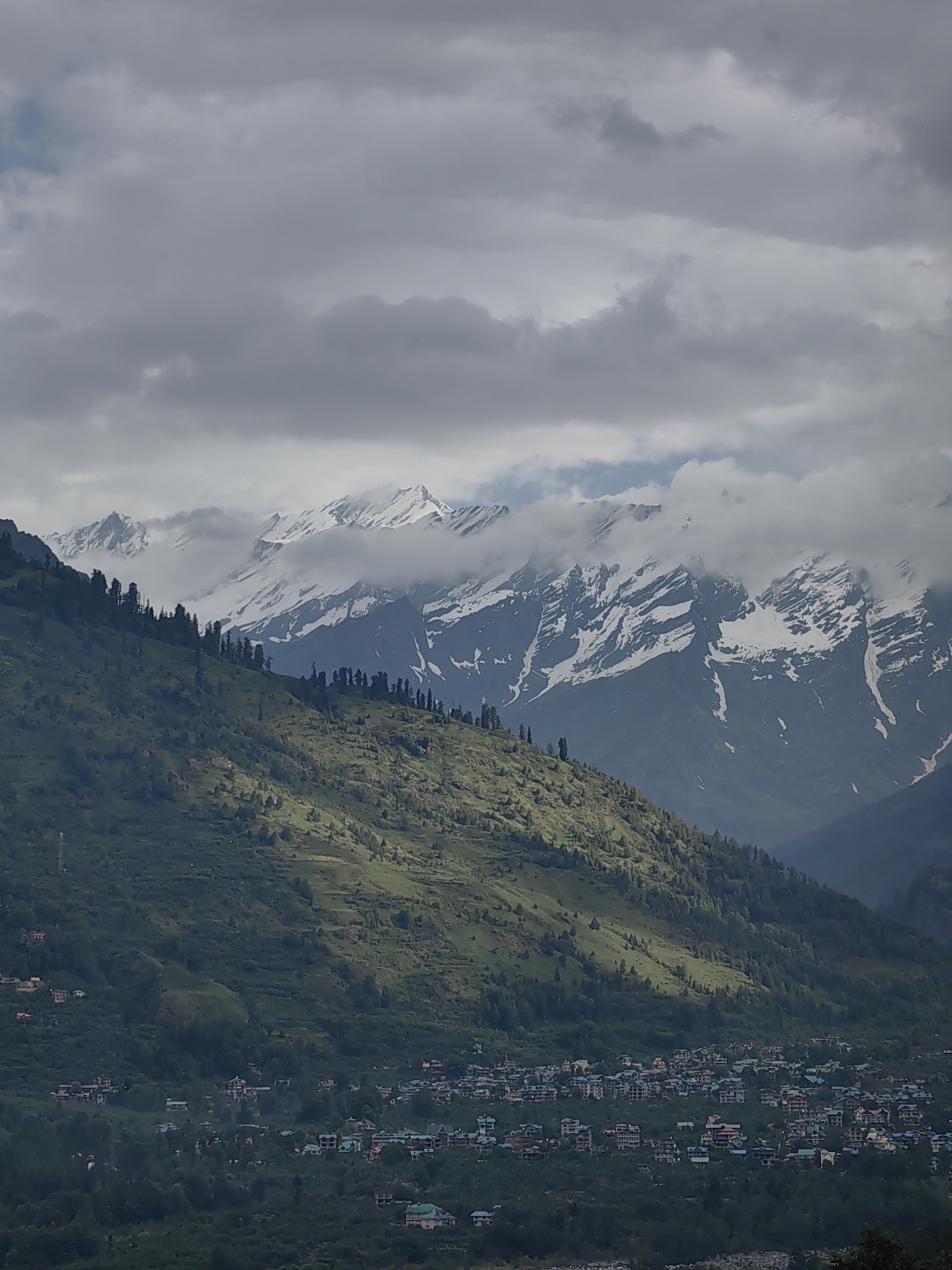 This is a vertical, medium-shot photograph of a mountainous landscape with a village in the foreground. The sky is overcast with thick, grey clouds that partially obscure the snow-capped peaks of distant mountains. The mountains themselves are rugged and majestic, with patches of snow clinging to their upper reaches. In the mid-ground, a verdant green mountain slopes downward, dotted with scattered trees. A cluster of buildings, indicative of a village or town, is nestled in the valley below the slope, surrounded by dense foliage. The buildings are low-rise structures, some with colorful roofs, and appear to be residential or commercial establishments. The overall scene conveys a sense of natural beauty and a remote, possibly rural, setting. Given the distinctive terraced slopes and the type of architecture, the location is likely Vashisht, India, a popular tourist destination known for its hot springs and proximity to the Himalayas. There are no people visible in the frame. The lighting is diffused due to the cloud cover, creating soft shadows on the landscape.