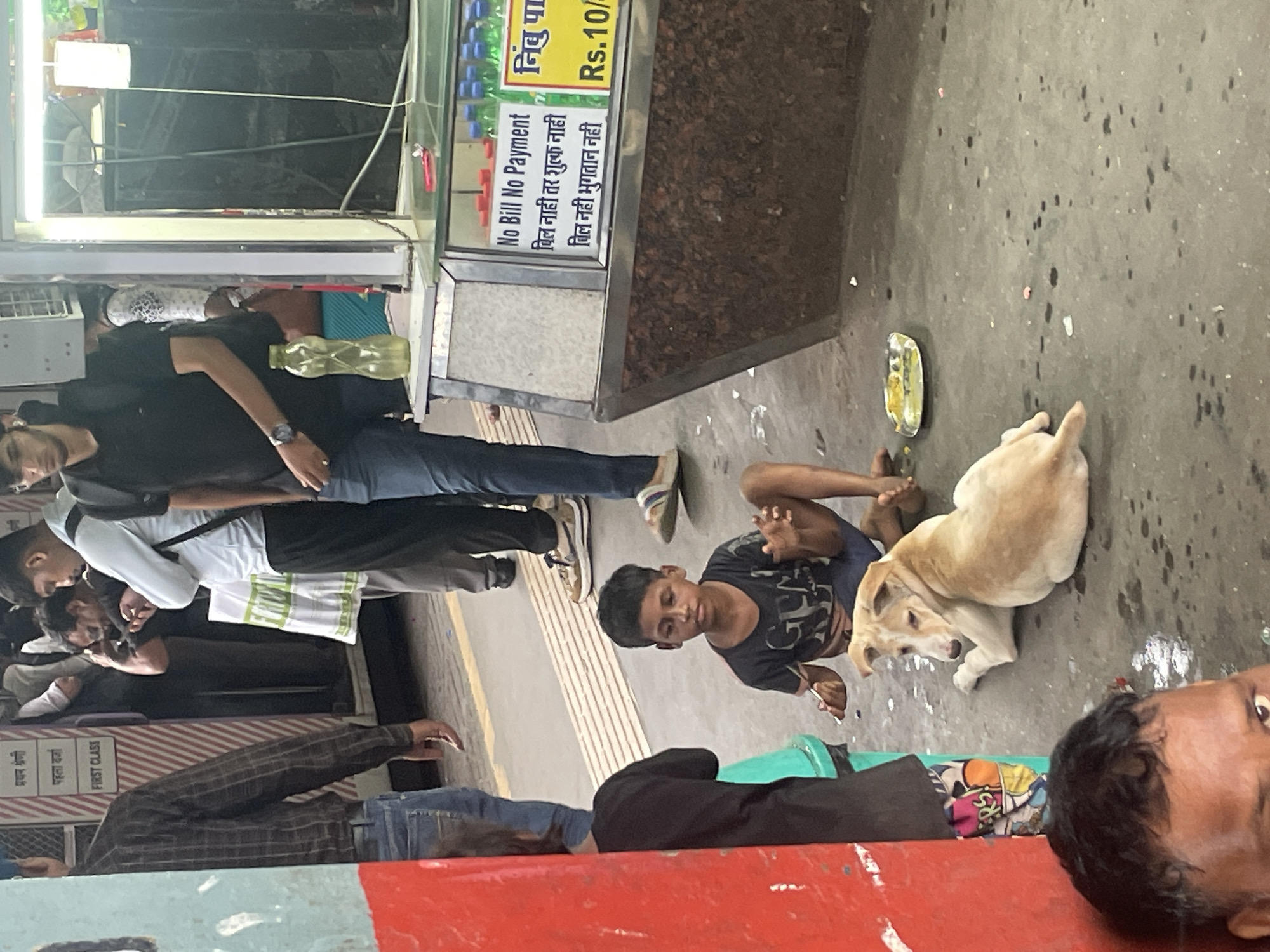 The image depicts a bustling scene at what appears to be a Mumbai railway station, possibly Dadar or Chhatrapati Shivaji Maharaj Terminus, judging by the architecture and signage suggesting "FIRST CLASS" compartments. In the foreground, a young boy, likely a street child, sits on the ground wearing a dark t-shirt and shorts. He is partially feeding a light-colored dog that is lying down next to him, looking up at him. A small metallic tray with some food is placed on the ground in front of the dog.
Behind the boy, several individuals are visible. A man in a blue shirt and jeans is walking away, his back to the camera. To his left, other people are standing, some looking at their phones. A stall with various bottles of drinks and signage is to the right of the frame, with one sign clearly stating "No Bill No Payment" in English and Hindi.
The environment is the concrete platform of the train station, with some discarded debris and water stains on the ground. The overall atmosphere suggests a busy public space with diverse socioeconomic elements coexisting.