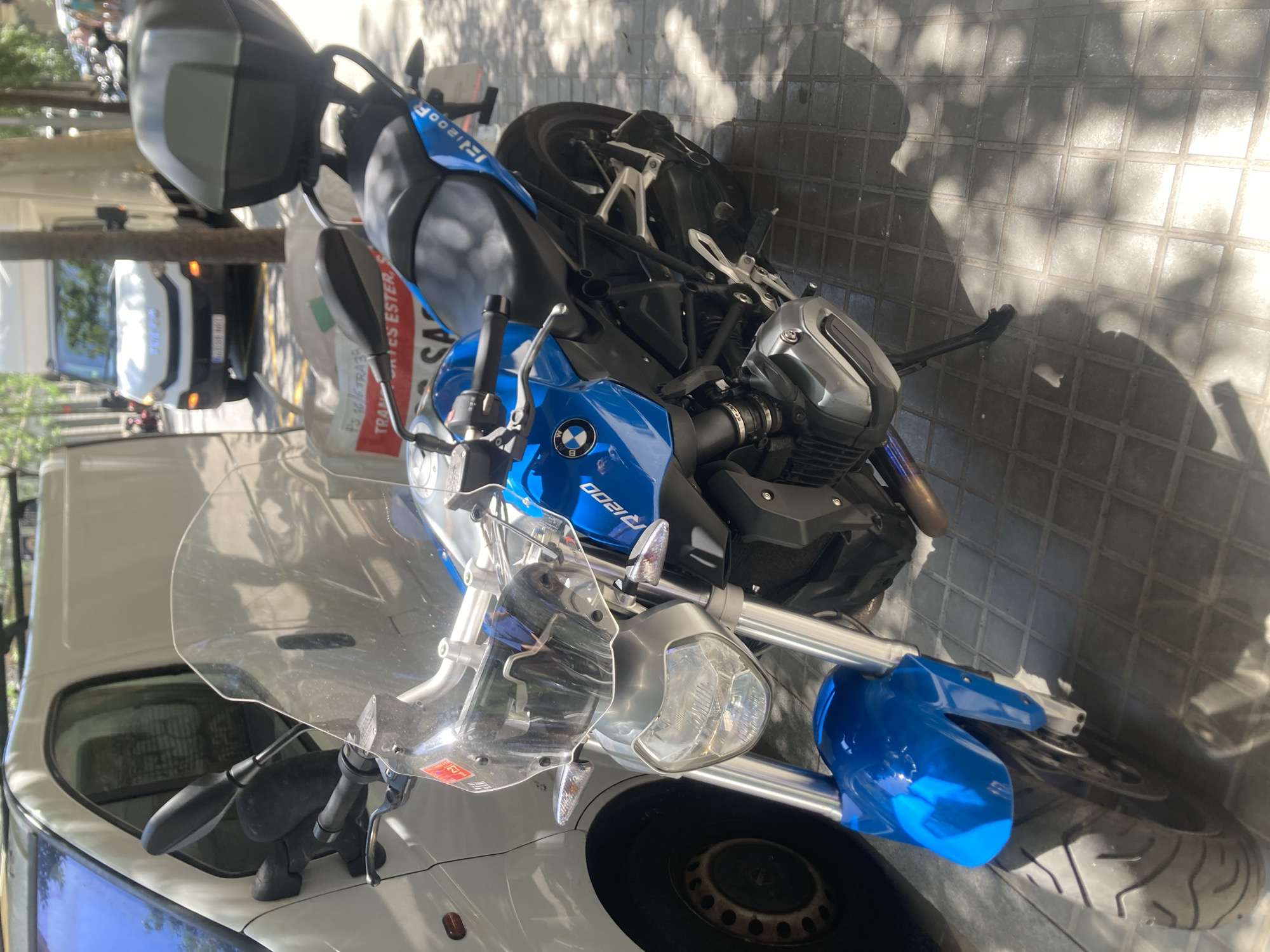 A blue BMW R1200 motorcycle is parked on a tiled sidewalk next to a white van in Barcelona, Spain. The motorcycle has a clear windshield, a black seat, and a gray storage box mounted on the rear. A white delivery truck is visible in the background, along with trees and buildings. The scene appears to be a street in a commercial or residential area.