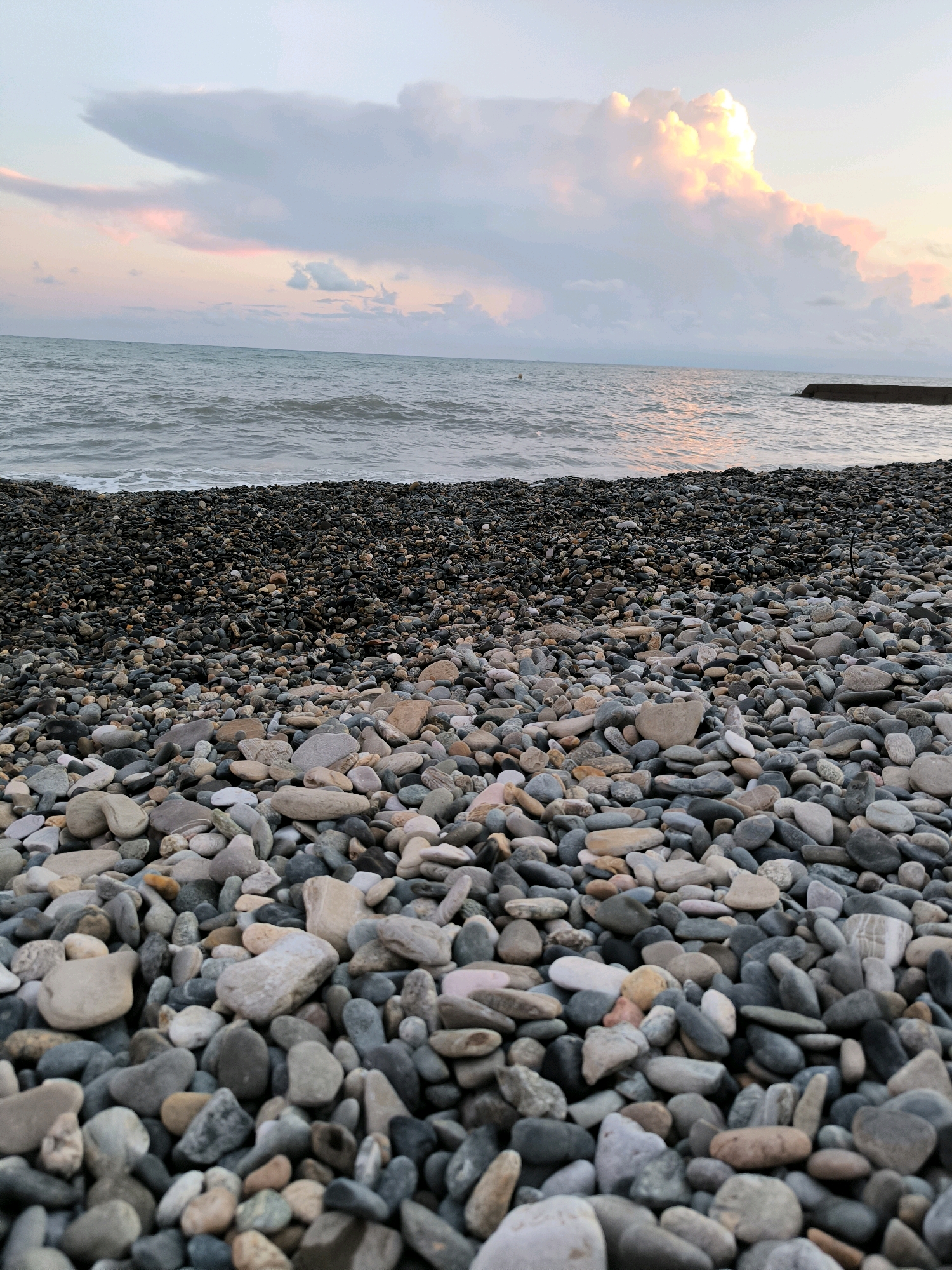 The image shows a pebble beach on the coast of Sochi, Russia. The foreground is filled with a variety of smooth, multi-colored pebbles of different sizes. Beyond the beach, the dark, choppy waters of the sea stretch to the horizon. The sky above is filled with dramatic clouds, illuminated by the soft, warm light of a sunset or sunrise. Some of the clouds are tinged with pink and orange, while others are a pale gray. In the distance, a concrete pier juts out into the sea from the right side of the frame, and a buoy is visible in the water on the left. The overall atmosphere is serene and natural, capturing the beauty of a coastal landscape at twilight.