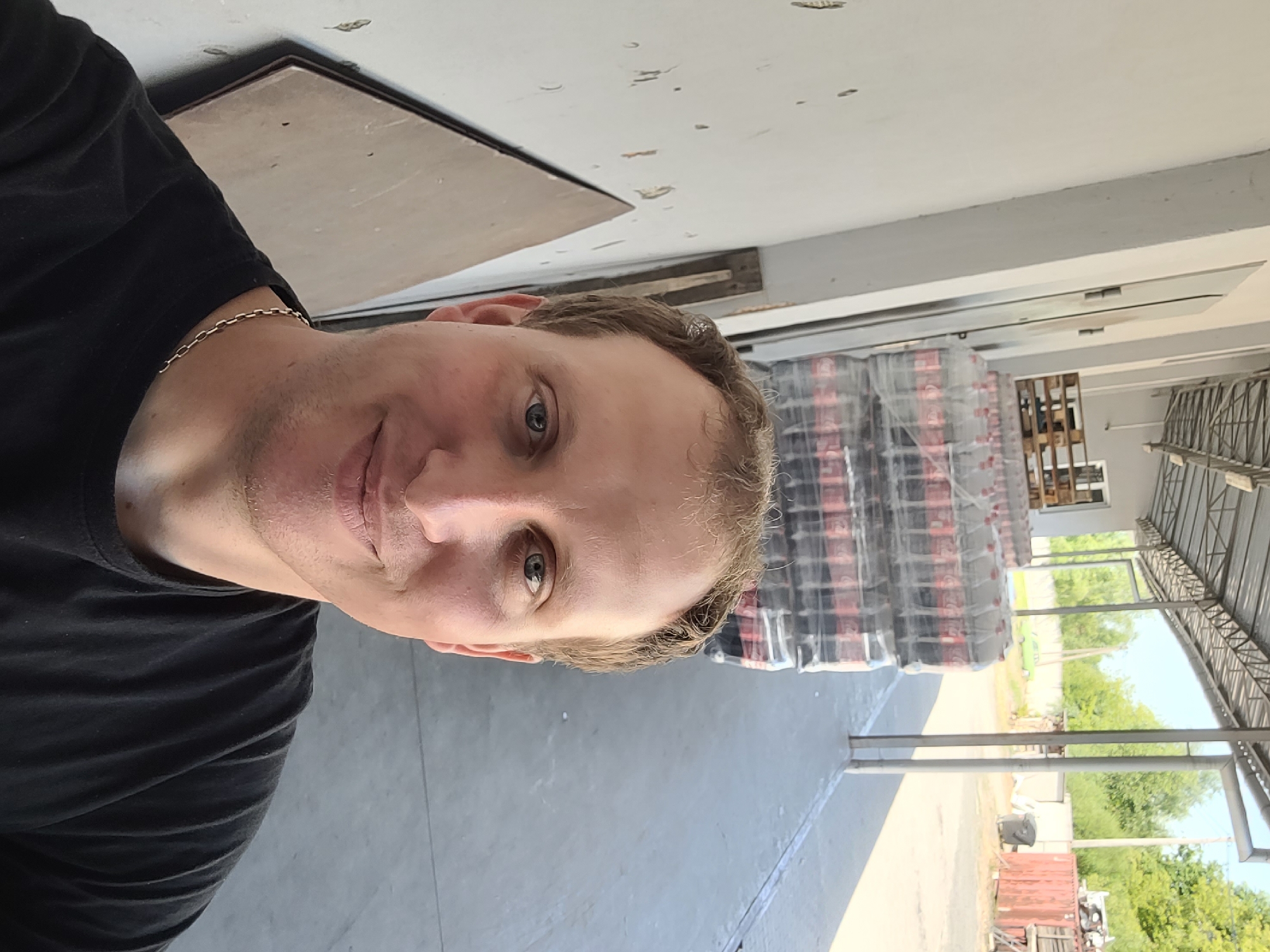 A Caucasian male with fair skin and short, light brown hair takes a selfie outdoors. He is wearing a black t-shirt and a silver chain necklace. He is smiling and looking directly at the camera. In the background, there are stacks of bottled drinks, pallets, a white wall, a covered walkway, and some trees. The ground appears to be paved. The lighting suggests it is daytime.