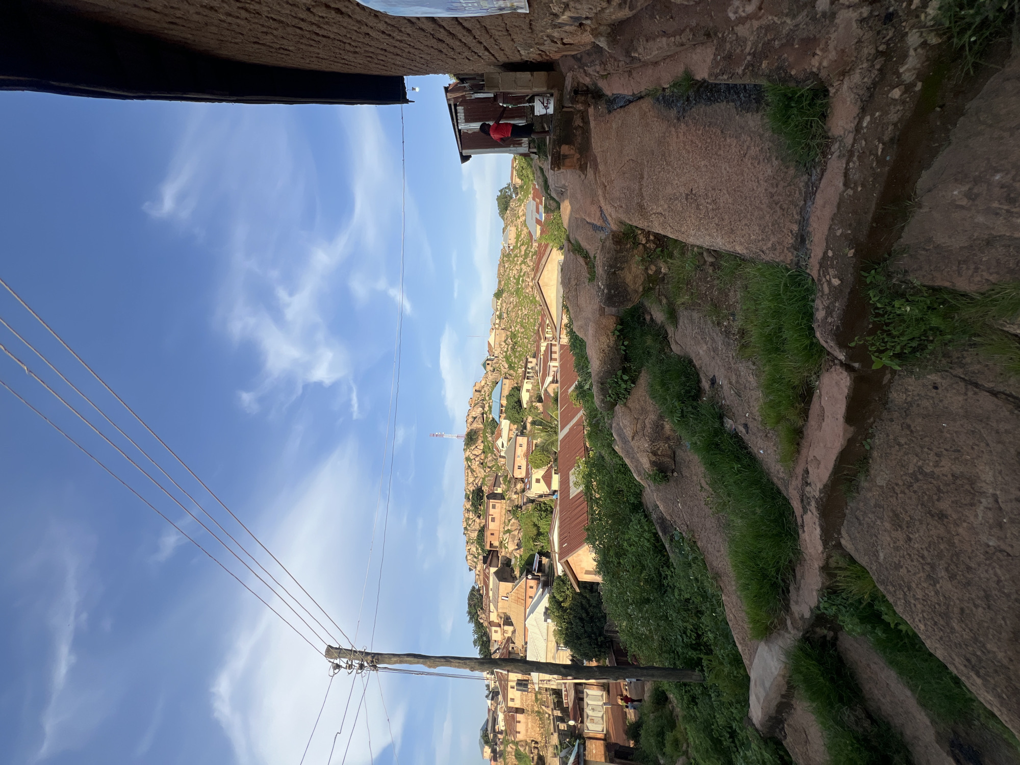 This image captures a daytime view of a residential area on a rocky hillside in the city of Jos, Nigeria. In the foreground, a rough, stepped path made of large, reddish-brown rocks and cement leads upwards and to the right. Green grass grows in patches between the rocks and along the edges of the path. To the right, the textured, mud-brick wall of a building dominates the immediate foreground, with a small corrugated metal structure attached to it. A young person wearing a red shirt and dark shorts is visible near the metal structure, standing by a water tap and a white bucket, appearing to be using the tap. A poster is affixed to the mud-brick wall.
Further up the hillside, a dense collection of houses with various roof types, including corrugated metal and what appears to be tiled roofs, are scattered among the rocks. The buildings are predominantly made of earth or brick. Green vegetation, including trees and shrubs, is interspersed throughout the settlement. In the background, a prominent rocky outcrop forms the peak of the hill, with more structures visible on its slopes. A tall communications tower can be seen on the far right of the hill's summit. Utility poles and multiple power lines stretch across the sky in the mid-ground, originating from a pole on the left side of the image. The sky is a bright blue with scattered white clouds, suggesting clear weather. A few small figures are visible on a path between buildings in the mid-ground to the left. The overall scene depicts a densely populated, hilly urban environment with a rustic and natural character.