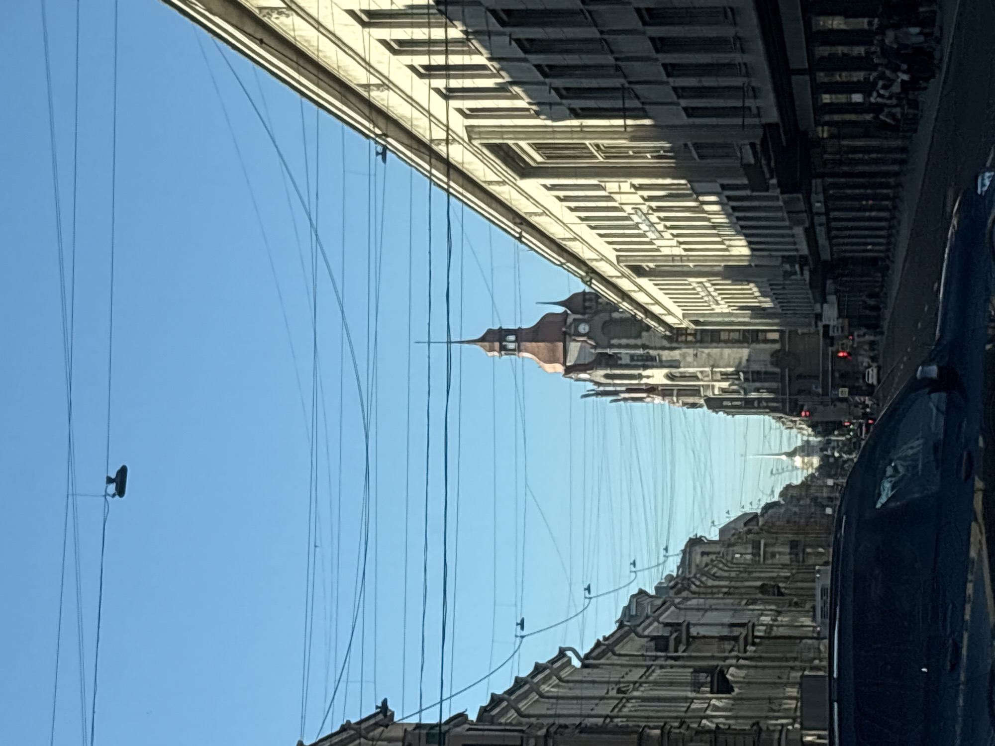 The image depicts a street scene in Saint Petersburg, Russia, likely on a clear, sunny day. The perspective is from a vehicle, looking down a street lined with architectural buildings.
On the right side of the street, a large, modern-looking building with a pale facade and numerous windows dominates the foreground. Its architecture suggests a commercial or administrative function, with the sun casting strong shadows across its surface. Further down the street, people are visible walking along the sidewalk, and some appear to be waiting or gathered.
On the left side, older, more ornate buildings with traditional St. Petersburg architecture are visible. These buildings have detailed facades and are also casting shadows.
In the middle distance, a distinctive tower with a red roof and a clock face stands out. Further down the street, another spire, possibly belonging to a church or another significant landmark, pierces the sky.
The sky is a clear blue, crisscrossed with numerous power or communication lines, a common sight in urban environments. In the upper portion of the image, a pair of shoes is suspended from one of these lines, a curious and often seen urban detail.
The overall impression is of a bustling city street with a mix of historical and modern architecture, under a bright sky, viewed from the perspective of a moving vehicle.