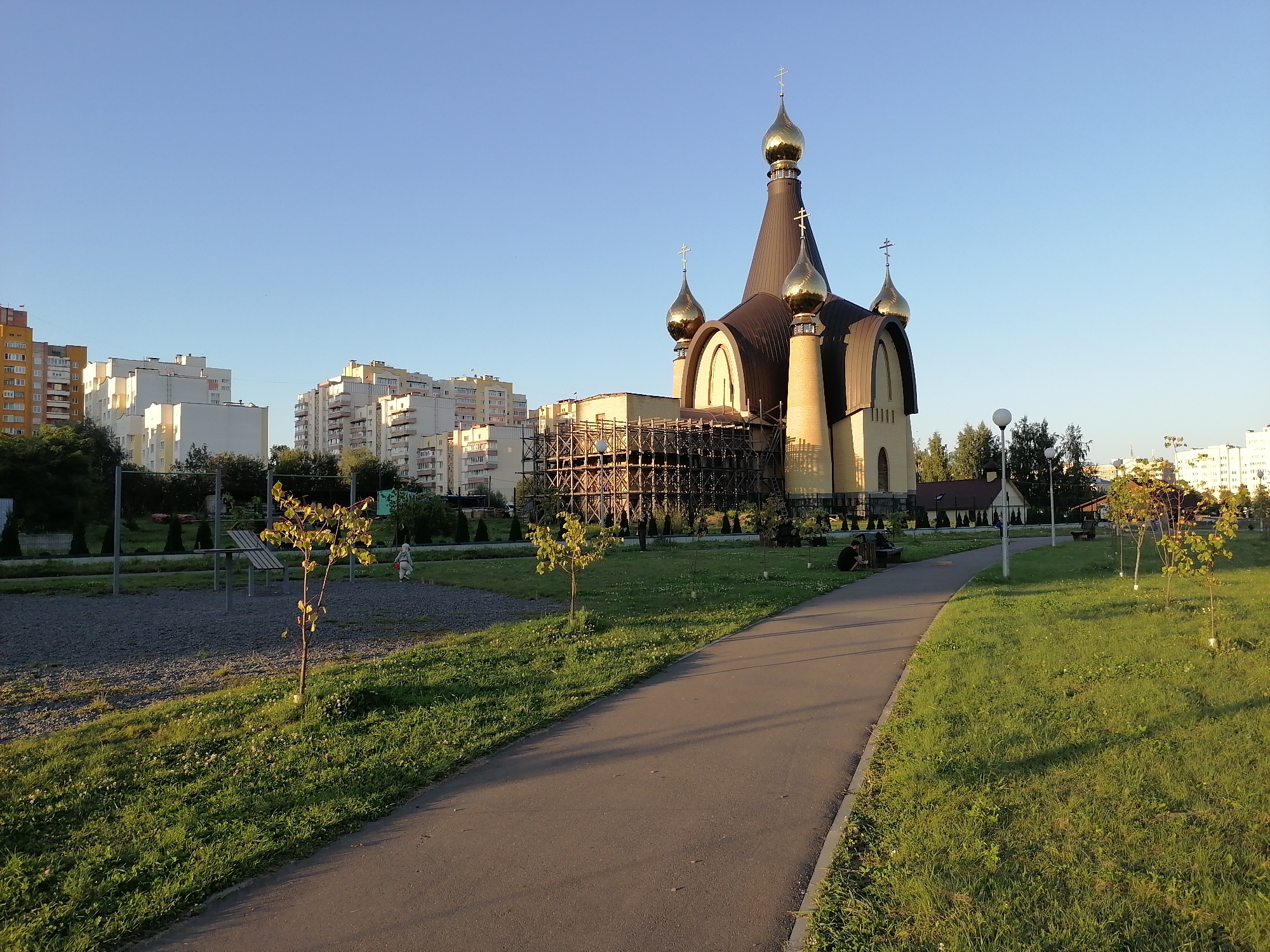 In the city of Hrodna, Belarus, a park area is pictured during daylight. A paved walkway runs diagonally from the lower left to the upper right of the frame, bordering a grassy area on the right and a gravel area with some young trees and playground equipment on the left. In the background, a domed church with golden accents is under construction, partially covered in scaffolding. Beyond the church, apartment buildings and trees are visible under a clear blue sky. A few people are present in the scene: one person is sitting on a bench in the park, and another is walking on the far left.