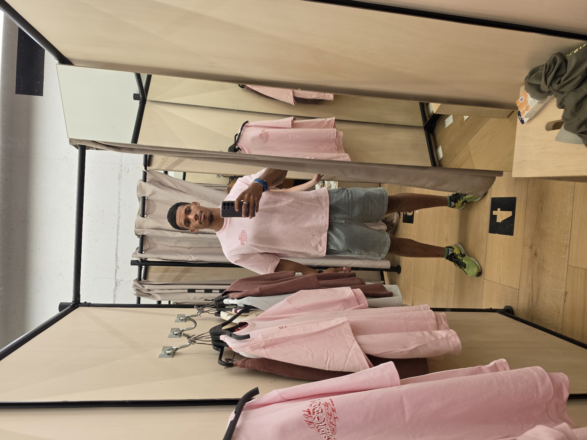 In this vertically oriented photograph, a young man stands in what appears to be a fitting room or retail space, taking a selfie in a mirror. He is wearing a light pink t-shirt with a graphic design and light blue denim shorts. His hair is short and dark, and he is looking at the camera with a neutral expression. He is holding a smartphone in his left hand, which is angled to capture the reflection.
The setting features multiple mirrors that reflect the man, the clothing racks, and the interior of the room. Various pieces of pink clothing are visible on hangers, some neatly folded on shelves and others displayed on racks. The walls are a light beige color, and the floor is made of light-colored wood. In the background, a beige curtain hangs, suggesting a changing area. To the right, a wooden surface holds a green hooded sweatshirt and a small carton of orange juice. The lighting appears to be artificial, typical of an indoor retail environment. The overall impression is a casual snapshot of someone trying on clothes.