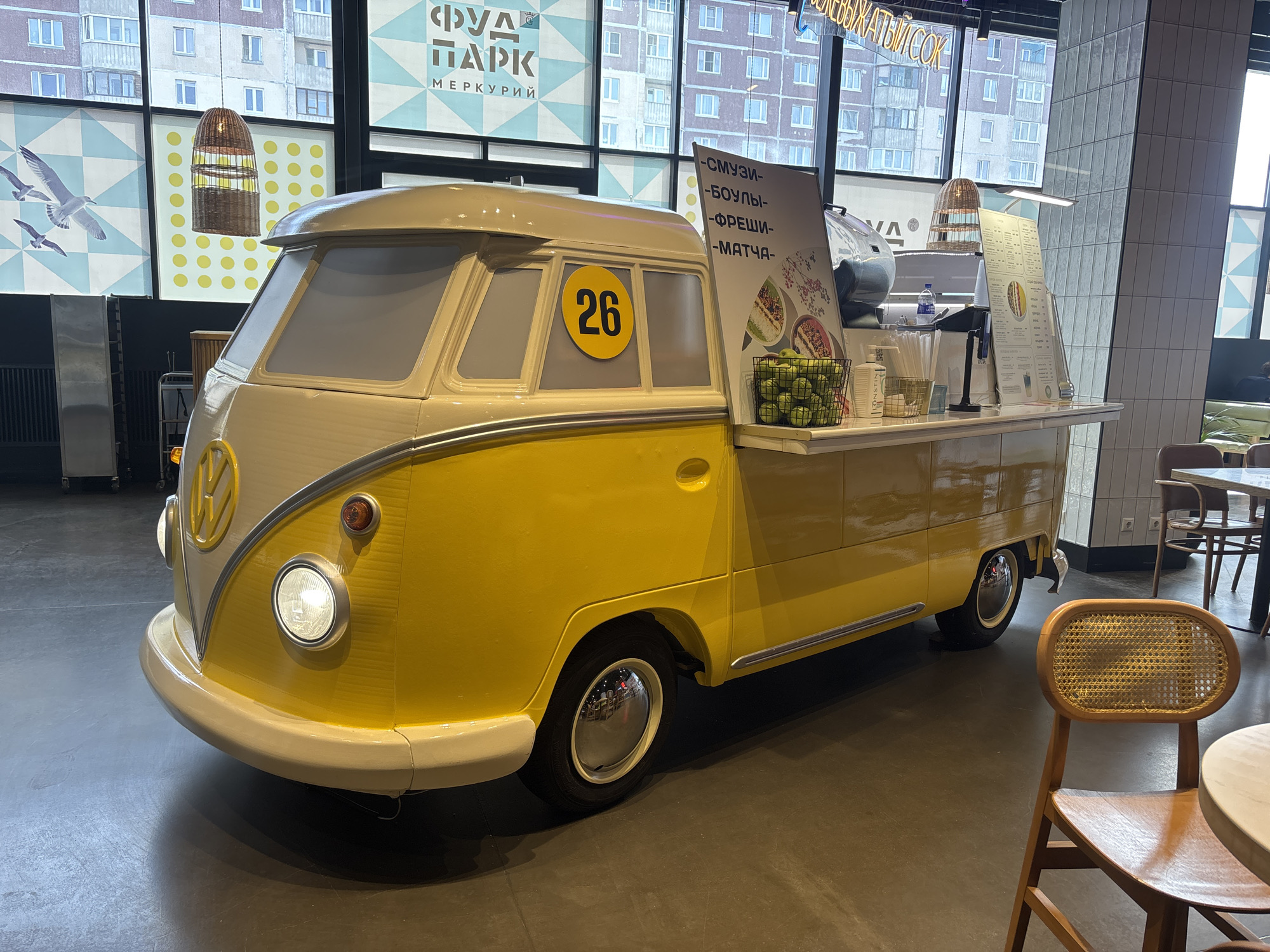 A yellow and cream Volkswagen bus, modified to serve as a food kiosk, is indoors on a gray, polished floor. The front of the bus is yellow and cream with a round "VW" logo and round headlights, one of which is illuminated. A yellow circle with the number "26" is painted on the side of the bus. On the bus's modified flatbed are a menu board listing "СМУЗИ, БОУЛЫ, ФРЕШИ, МАТЧА", and a basket of green apples. A wooden chair is in the foreground. In the background, a large window provides a view of an apartment building and a food park sign reading "ФУД ПАРК МЕРКУРИЙ" above the bus. This photograph was taken in Saint Petersburg, Russia.