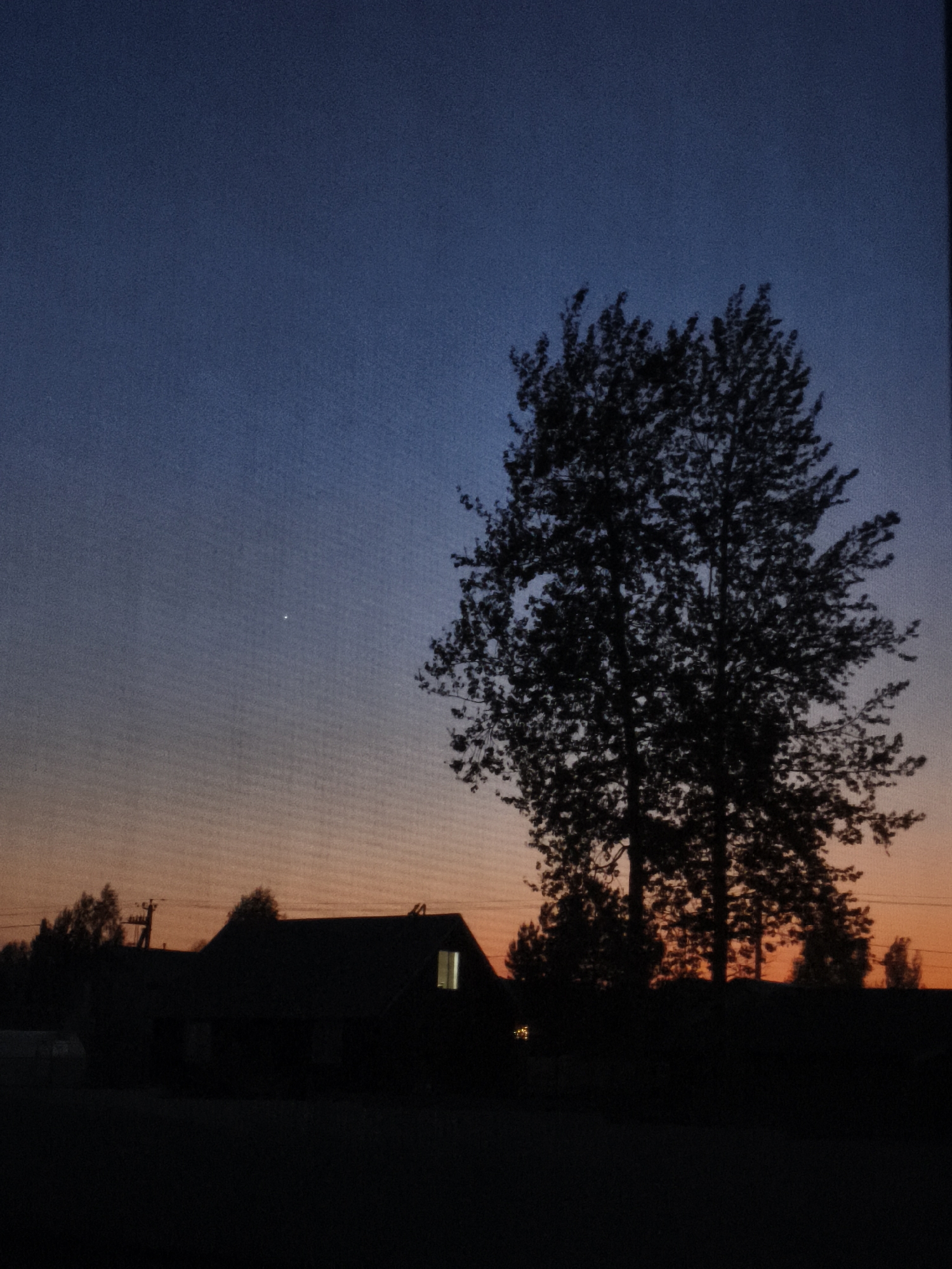 The image depicts a twilight scene in Русский Вожой, Russia, dominated by a silhouette of a building and a large tree against a gradient sky. The sky transitions from blue at the top to orange near the horizon. A single bright star or planet is visible in the upper left quadrant. The building is dark, but a window glows with interior light. The tree is leafless or has small leaves, obscuring the background. The foreground is dark, suggesting a ground-level view from a window. Other silhouetted trees or structures are visible on the horizon behind the building. There are no visible people, actions, or interactions.