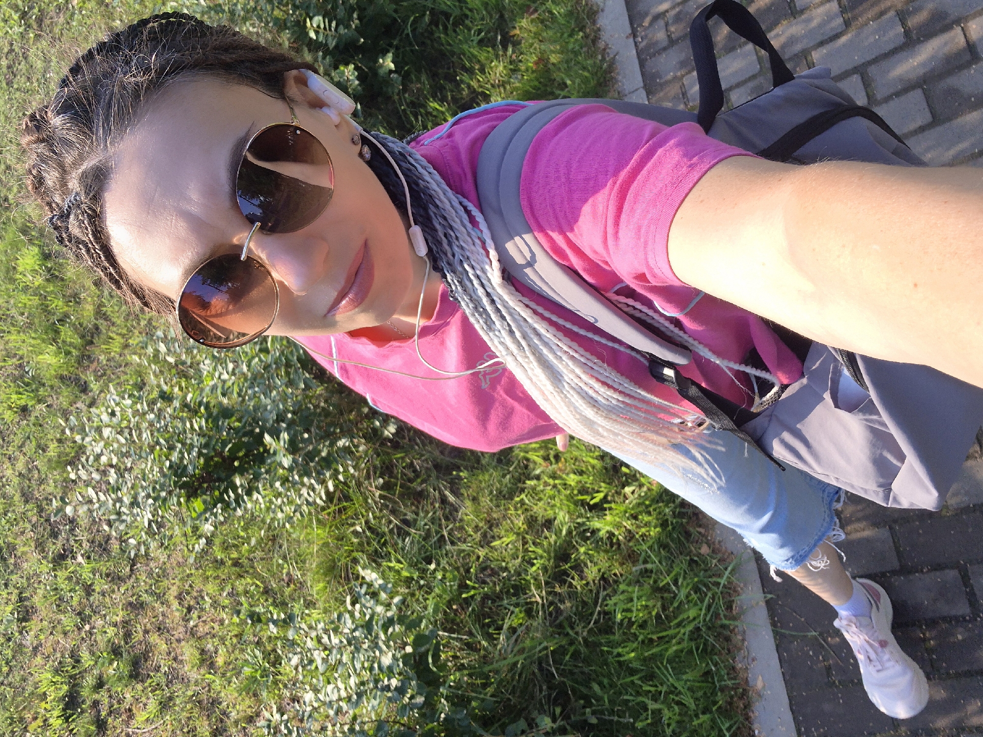 A woman with braided hair is taking a selfie outdoors. She is wearing a bright pink t-shirt, light blue ripped jeans, white sneakers, and round sunglasses. White earbuds are in her ears. She is also wearing a grey backpack. The background consists of green grass and a paved walkway. The lighting suggests it is daytime. The image is likely from Chaikovsky, Russia.