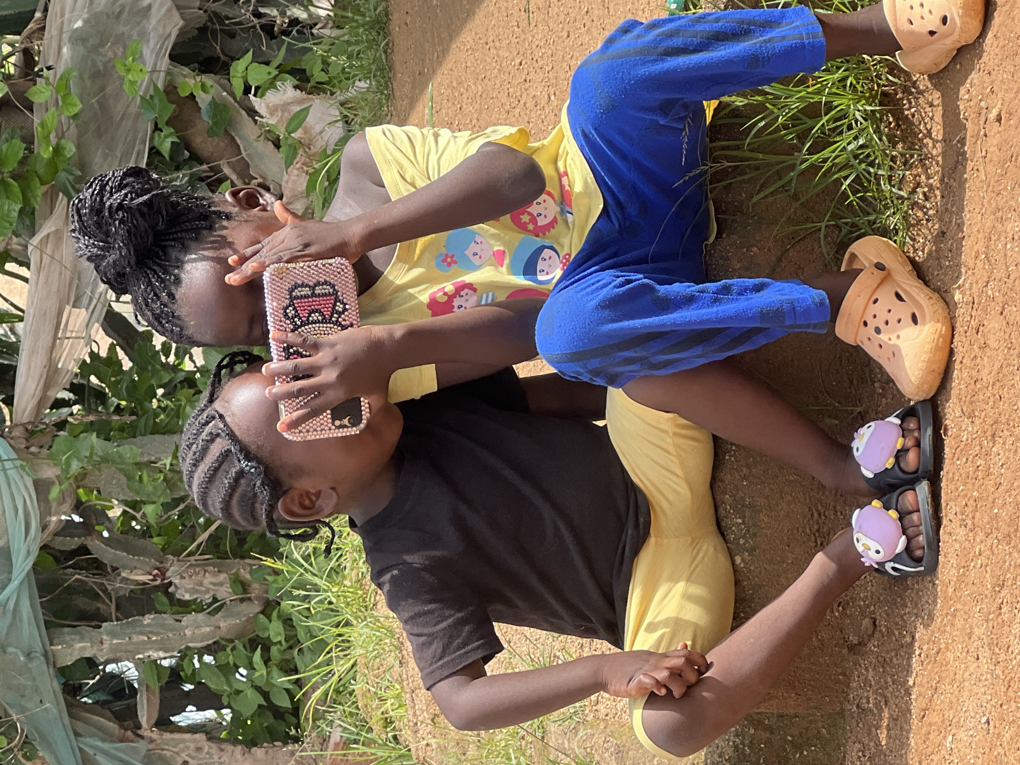 Two young children, a boy and a girl, are sitting on a concrete ledge outdoors. The boy, on the left, wears a black t-shirt and yellow shorts. He is holding a pink phone case adorned with rhinestones up to his face, looking at it with curiosity. The girl, on the right, is wearing a yellow t-shirt with cartoon characters and blue striped pants. She is also holding a pink phone case, similar to the boy's, and appears to be using it. Both children have their hair braided. They are barefoot, with the boy wearing black slides decorated with purple owl charms and the girl wearing light orange Croc-style shoes. The background consists of dry vegetation, green foliage, and what appears to be some sort of covering or netting. The ground is a reddish-brown dirt. The scene suggests a casual, outdoor setting, potentially in a residential area of Jos, Nigeria, given the dry climate and common vegetation.