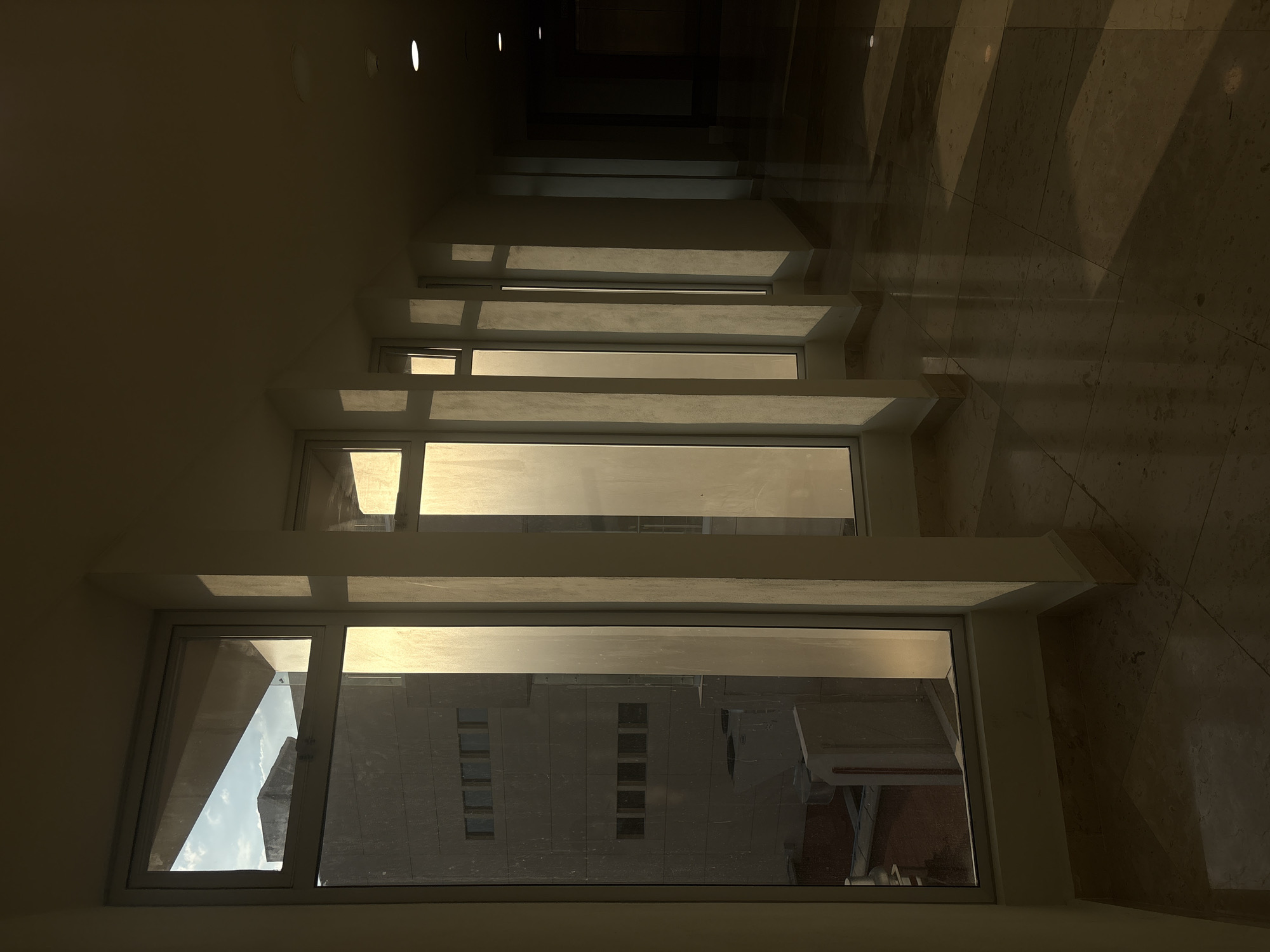 A series of windows viewed from inside a building. The windows descend diagonally into the distance, with buildings visible through the glass. The interior walls are light-colored, while the floor appears to be marble or a similar polished stone, reflecting light. Light patterns on the floor suggest sunlight streaming into the space. The location is Puebla, Mexico, based on the architecture of the buildings visible through the windows.