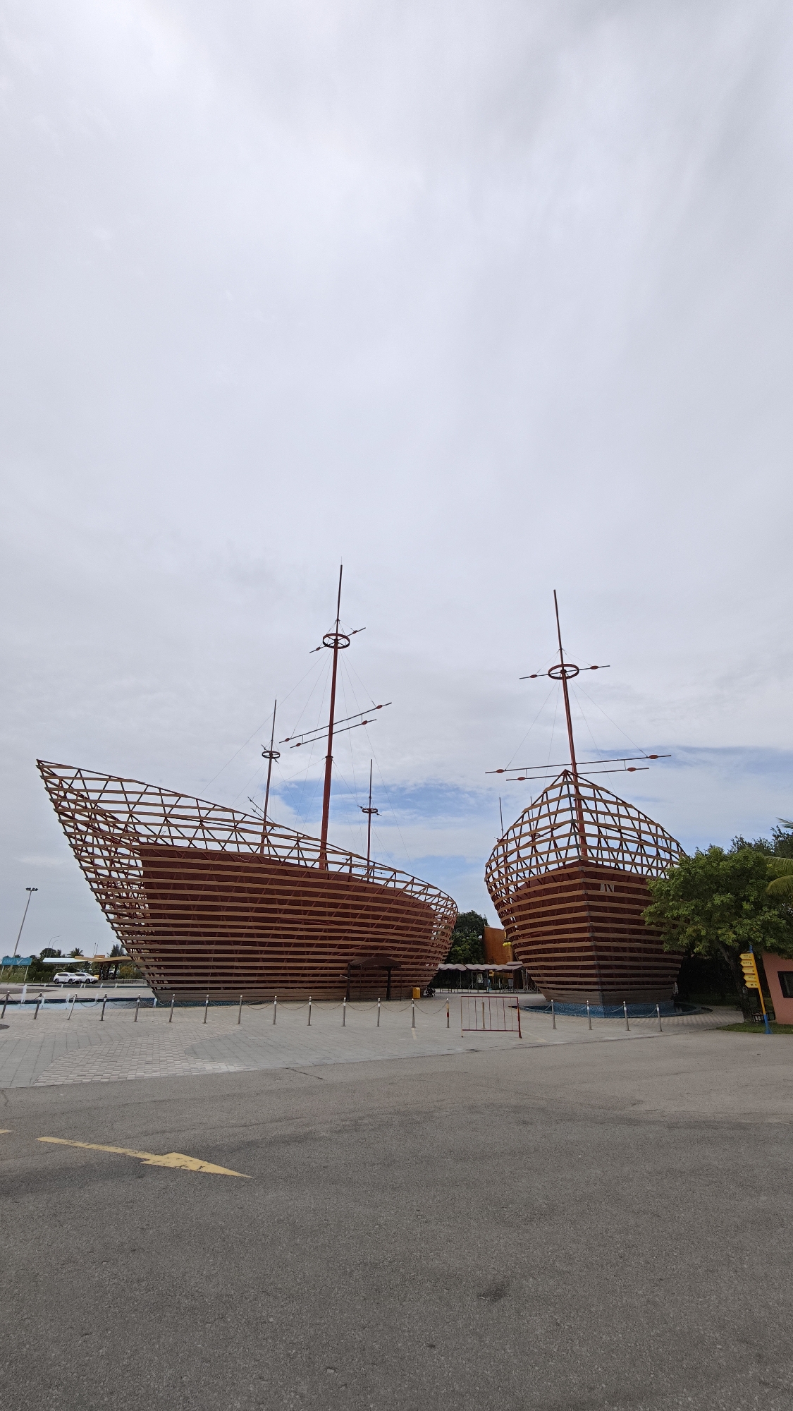 Two large, non-operational wooden ship structures are positioned side-by-side in an open, paved area. Each ship has a wooden frame hull with horizontal planking and masts with crossbars. The ships are likely decorative or representational rather than functional vessels. A yellow directional arrow is painted on the ground in the foreground. The sky is overcast with a mix of light and dark clouds. Trees and buildings are visible in the background. The structures are located in Đà Nẵng, Vietnam.