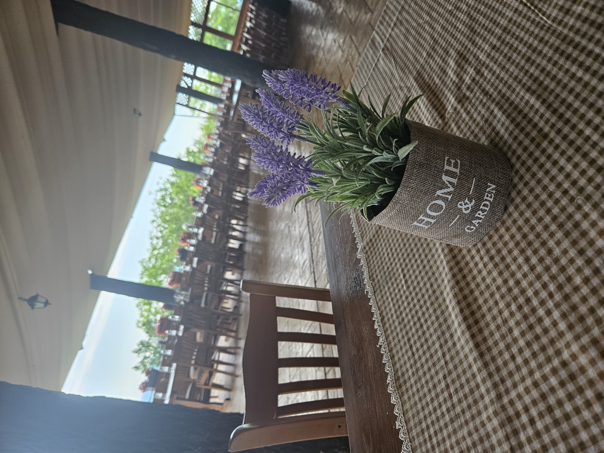 The image depicts an outdoor dining area with a rustic ambiance, set in the city of Lyaskovets, Bulgaria. In the foreground, a wooden table is covered with a brown and white checkered tablecloth, adorned with a decorative lace trim. A small burlap planter holds artificial lavender, bearing the inscription "HOME & GARDEN." Behind the table, a wooden chair is visible. The background reveals a long row of wooden tables and chairs, suggesting a restaurant or event space. The area is covered by a large, beige canopy, supported by dark wooden pillars. Beyond the canopy, lush green trees and a bright sky indicate a natural setting. The flooring appears to be tiled or paved. The overall scene conveys a peaceful, outdoor dining experience in Lyaskovets.