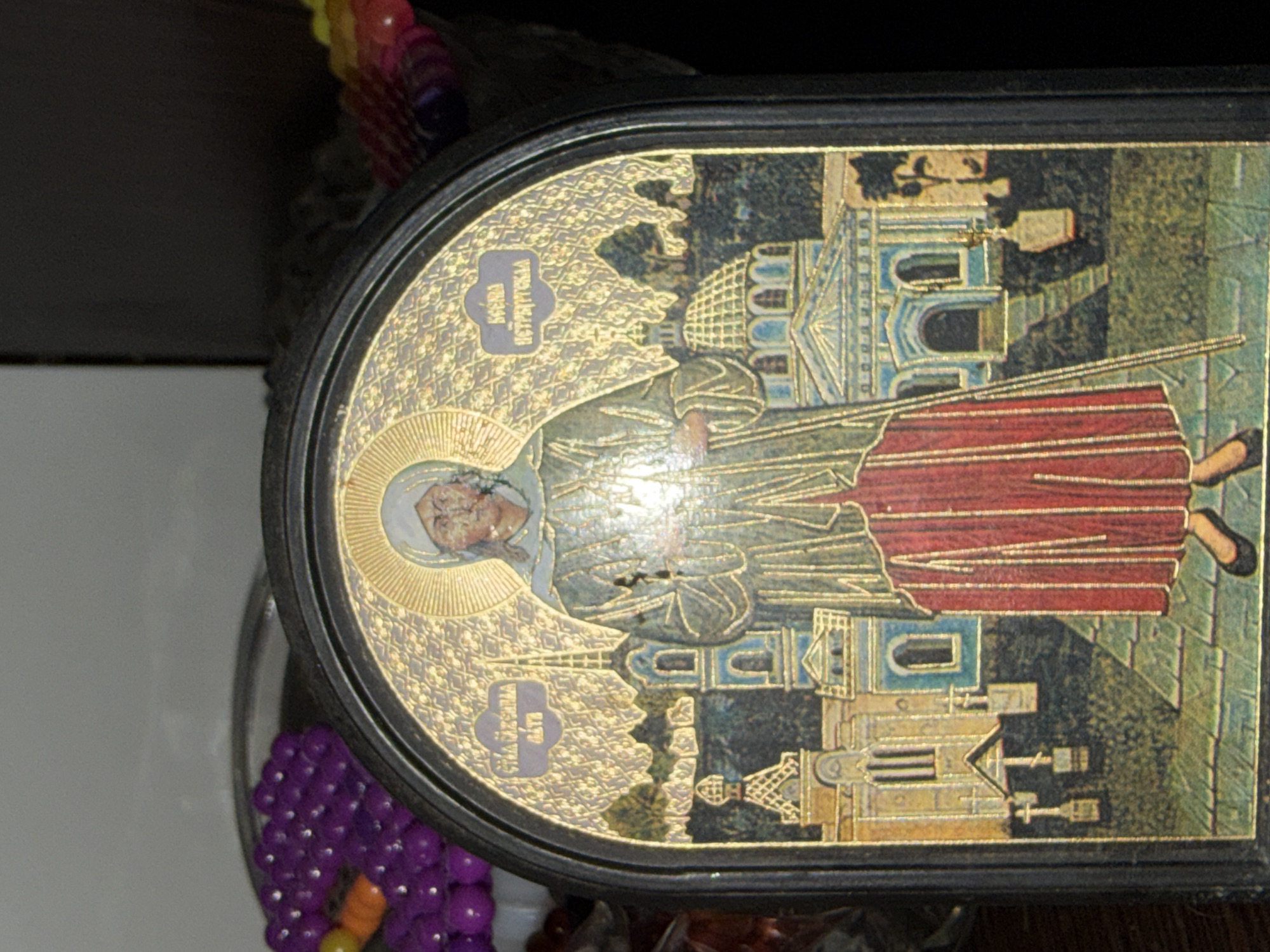 The image is a close-up of an Eastern Orthodox icon. The icon depicts a female saint with a halo, dressed in traditional religious garments, standing before a cityscape featuring churches and buildings. The artwork is characterized by detailed patterns and a gilded background. Inscriptions in a Cyrillic script are visible on the icon. The overall impression is of a religious artifact with an aged appearance, suggesting it is an antique or religious relic. The image does not provide any information or context related to the city of Shepsi, Russia.