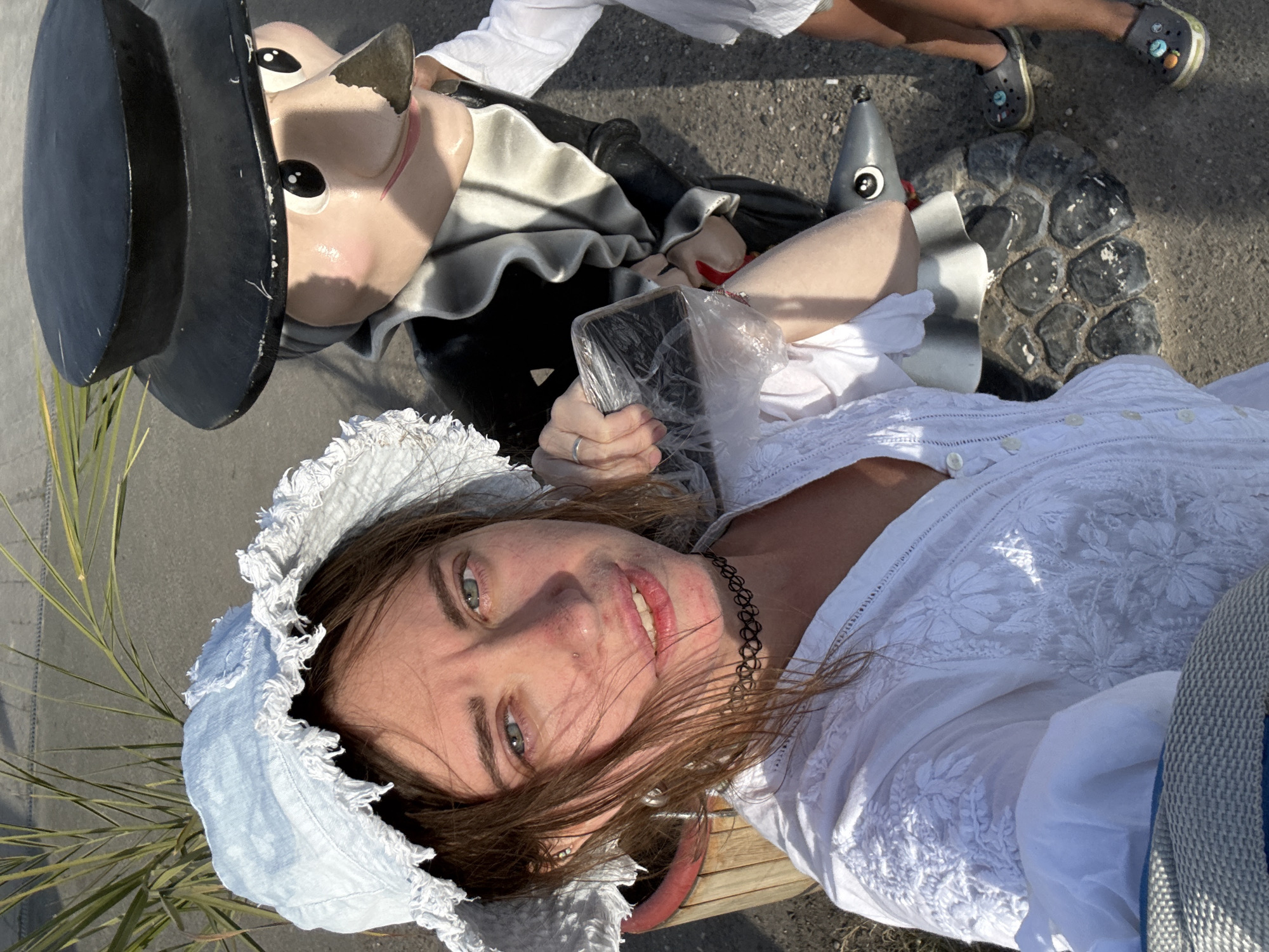 A woman is taking a selfie with a cartoonish statue of a man in a black hat, white frilly collar, and black suit. The woman is wearing a white embroidered dress and a light blue bonnet with a ruffled brim. She has brown hair and blue eyes and is wearing a black choker necklace. She is holding a clear plastic-wrapped object in her right hand. To the right of the statue, a child's legs and feet are visible wearing dark crocs. The statue is standing on a cobblestone base. The background shows a paved surface, possibly a sidewalk or street, and a palm frond from a nearby plant. The scene appears to be outdoors, under bright sunlight. The statue is located in the city of Sol-Iletsk, Russia, possibly at a tourist attraction or public space.