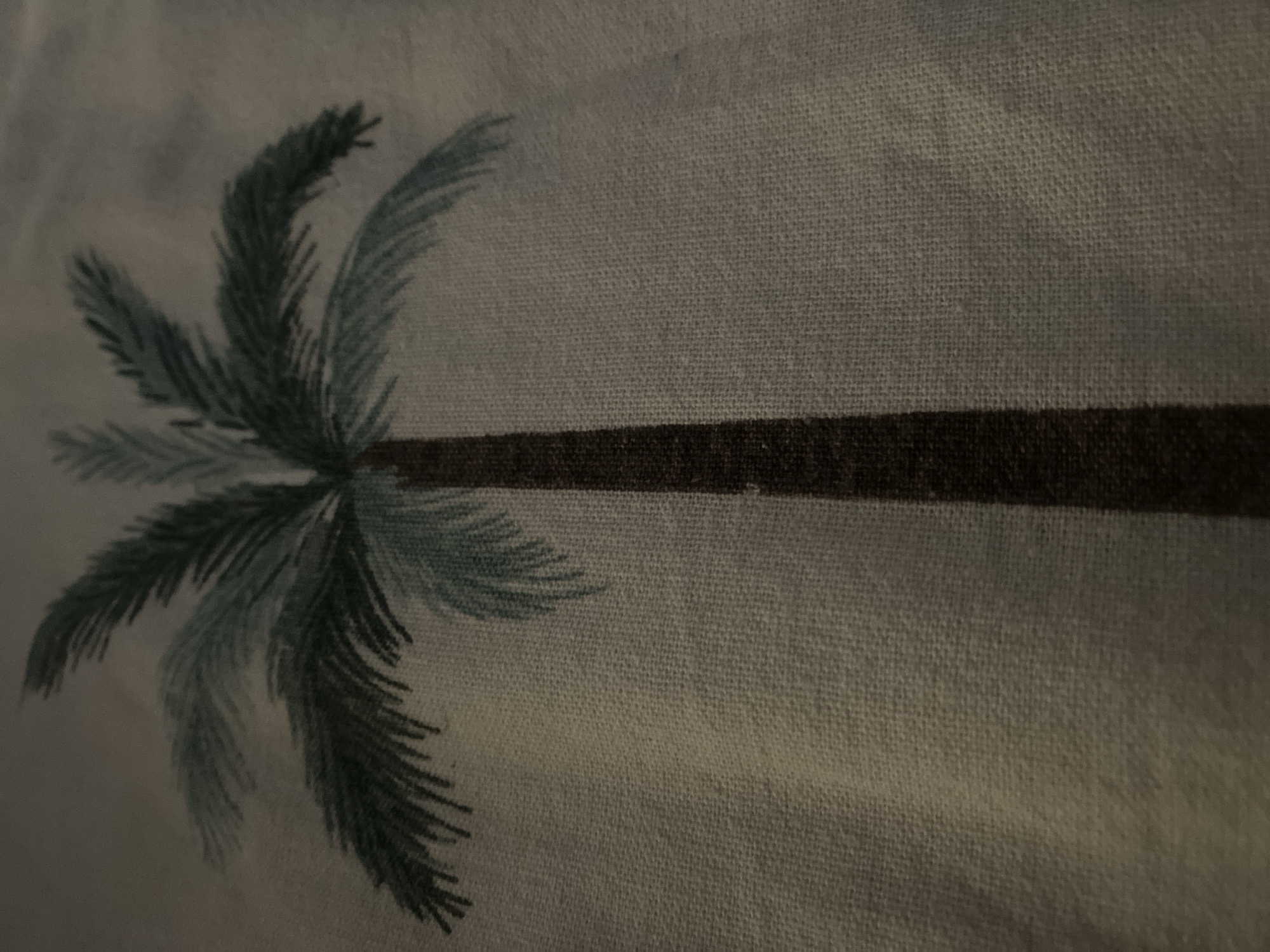 A close-up shot displays a stylized painting of a palm tree. The trunk of the tree is a dark brown, and its fronds are rendered in varying shades of dark green and a lighter blue-green. The background is a muted, light gray, suggesting fabric or a textured surface. The image lacks any details of a specific city or location.