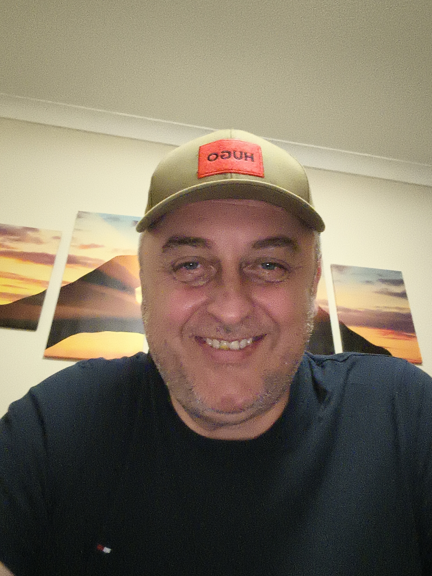 A close-up selfie captures a smiling middle-aged man indoors, likely in Huntingdonshire, United Kingdom. He is Caucasian, with short, greying hair visible at the sides and stubble on his chin. His light-colored eyes are open wide, and he displays a broad, genuine smile, showing his teeth. He wears a khaki-green baseball cap with a prominent red patch on the front, featuring the mirrored text "OᗡUH" (intended to be "HUGO"). He is dressed in a dark blue or black t-shirt, which has a small, distinctive red, white, and blue rectangular logo on its lower left side, resembling the Tommy Hilfiger brand.
The man is positioned slightly below the camera, suggesting a selfie angle, and his expression conveys happiness and approachability. The background features a plain light-colored wall with a visible white ceiling line or coving at the top. Three rectangular canvases or framed prints adorn the wall behind him, depicting a vibrant sunset or sunrise scene over silhouetted mountains or hills, characterized by warm orange, yellow, and red hues in the sky and landscape. The image exhibits some digital noise or grain.