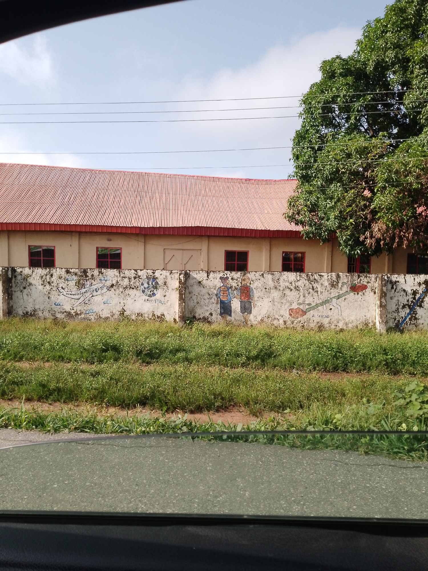 A long, low building with a corrugated red roof is visible. The building has cream-colored walls with several rectangular windows that have red frames. In front of the building is a rough concrete wall covered in a mural. The mural depicts various scenes, including an airplane, children playing on a seesaw, and a football. The wall is weathered and shows signs of age. A lush green field of grass and vegetation is in front of the wall. To the right of the building, a large, leafy green tree is prominent. The sky above is a pale blue with some scattered white clouds, and three horizontal power lines stretch across the scene. This image was taken from inside a vehicle, as evidenced by the reflection of the interior in the foreground. The location is Serti, Nigeria.