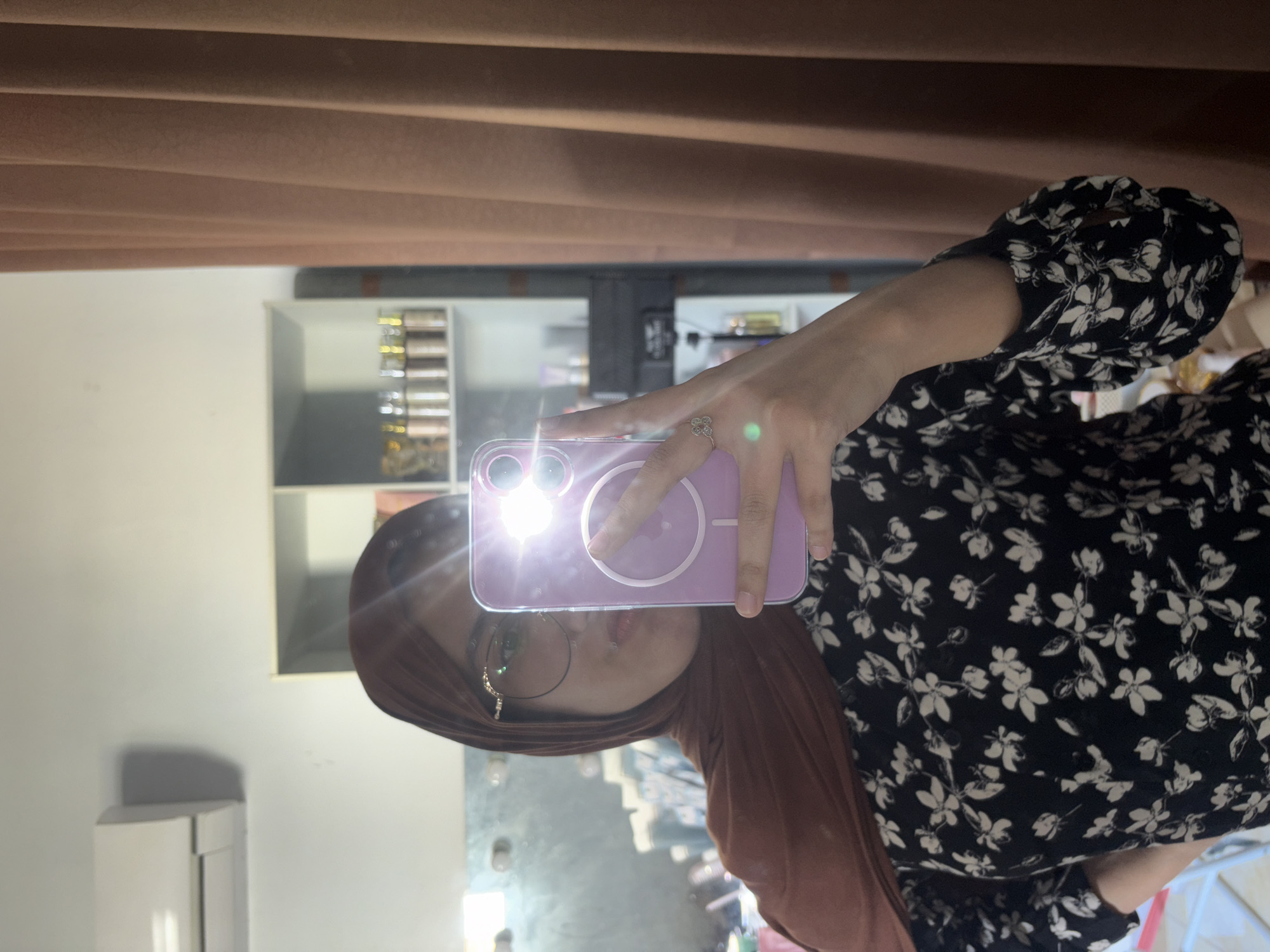 A person is taking a selfie in a mirror. The individual is wearing a hijab and glasses. The person is holding a purple phone with a flash turned on. The background includes a shelf with various items, a white wall, and a brown curtain. The location is presumably a personal space, possibly in Gadingrejo, Indonesia.