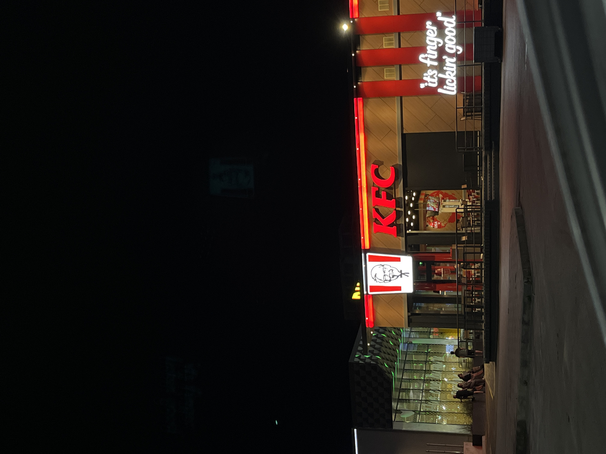 The image shows a KFC restaurant in Shumen, Bulgaria, at night. The building has a modern design with a large illuminated KFC logo and the slogan "it's finger lickin' good" in neon lights. There are several people sitting outside on a bench, and a few tables and chairs are visible in front of the restaurant. The surrounding area appears to be a parking lot or plaza. The overall scene is well-lit by the restaurant's signage and some ambient lighting from nearby structures.