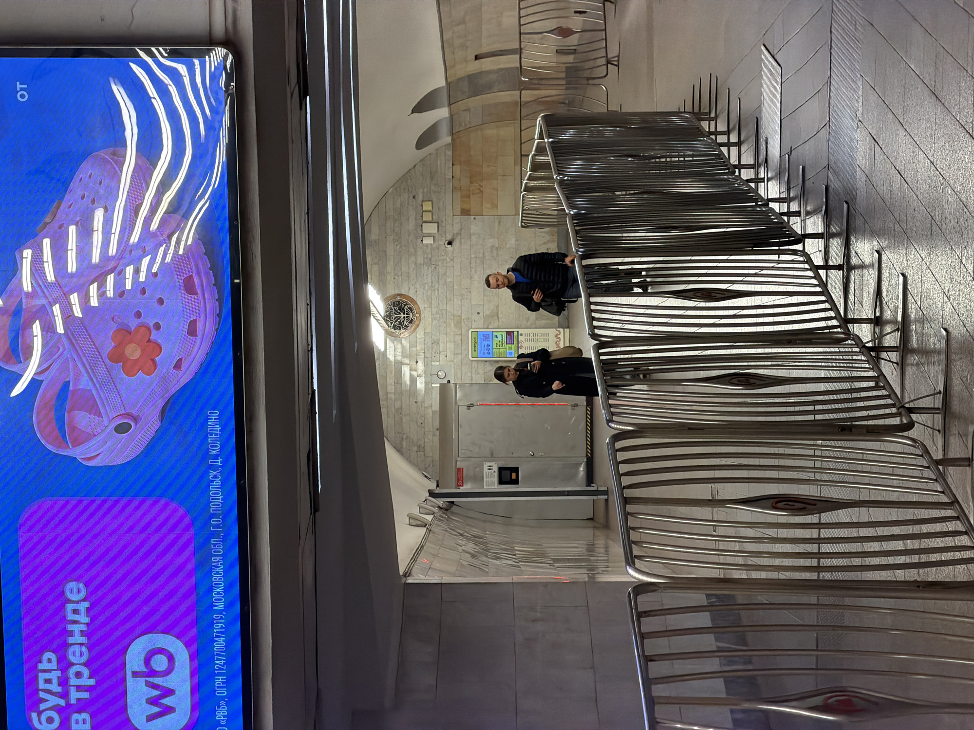The image captures a scene in what appears to be a subway station in Moscow, Russia. The perspective is unique, shooting from a low angle looking up and through metal barriers, providing a view into the station's interior. Two individuals are visible: one is a man wearing a blue shirt and a black jacket. The other appears to be a woman, dressed in black, with her back turned towards the camera. They are standing near what seems to be an automated teller machine (ATM).
The backdrop reveals the station's architecture, characterized by tiled walls, arched ceilings, and decorative elements. A circular ornamentation is visible on one of the walls. In the foreground, there is a vibrant advertisement on a screen featuring a pink sandal and a purple logo. It reads "Будь в тренде wb" (Be in trend wb) indicating that is a product of the Russian online retailer Wildberries (WB). The screen has text that reads: "OT", "РВБ, ОГРН 1247700471919, МОСКОВСКАЯ ОБЛ., Г.О. ПОДОЛЬСК, Д. КОЛЕДИНО".
The scene is a blend of commercial advertising, everyday transit, and architectural aesthetics, providing a snapshot of Moscow's urban landscape.