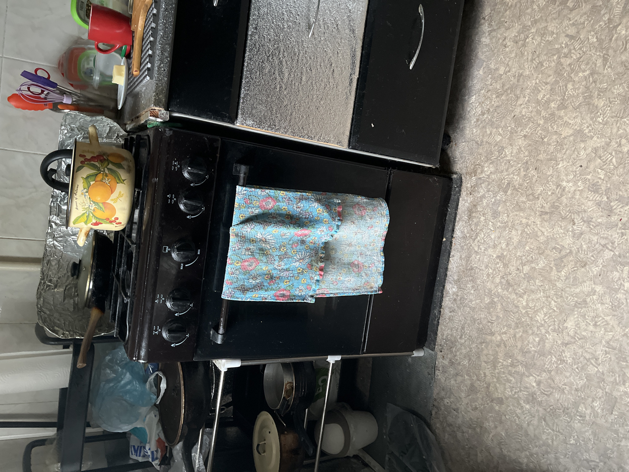 This is a forensic description of a kitchen scene located in Minsk, Belarus. The image captures a functional and lived-in kitchen space.
In the center of the frame is a black four-burner gas stove and oven unit. On the stovetop, a dark frying pan with a wooden handle rests on the back-left burner, while a decorative yellow enamel pot, adorned with oranges and berries and labeled "Aroma of Autumn," occupies the front-right burner. A light blue dishtowel with a floral pattern hangs from the black handle of the oven door. The stove's control panel features four black knobs with visible settings such as "MIN" and "MAX."
To the immediate right of the stove, a gray speckled countertop holds various kitchen items. A small white plate with a pat of butter, a red ceramic mug, and a collection of wooden cooking utensils are arranged on the surface. Adjacent to these, a clear utensil holder contains brightly colored plastic kitchen tools, including a whisk, tongs, and a spatula. A black textured mat is also present on the countertop. The wall above the counter is covered with white ceramic tiles. Below the countertop, a cabinet unit features an upper section covered with a textured silver foil-like material and a lower black drawer/door equipped with a curved silver handle.
To the left of the stove, a black metal shelving unit or utility cart stores additional kitchenware. A light blue plastic bag is visible on an upper shelf. Below, various pots and pans, including a dark pan with a lid, are stacked. Further down, a white bucket and other cooking vessels are stored on the bottom shelf. In the background, a light-colored wall is visible, with a roll of white paper towels and crumpled aluminum foil.
The floor throughout the kitchen is covered with light-toned linoleum or vinyl flooring featuring a dense, repeating, abstract pattern. No individuals are present in the scene, and no specific actions or interactions are depicted.