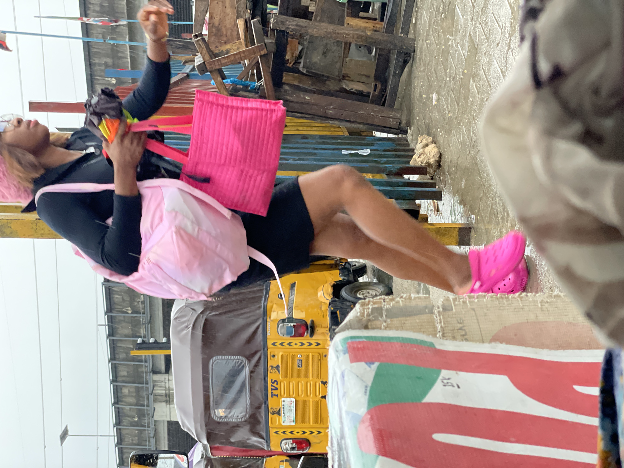 A woman wearing a black dress and pink crocs walks on a wet street in Lekki, Nigeria. She carries a large pink bag on her left shoulder and a smaller pink tote bag in her right hand, along with a folded umbrella. The street is lined with yellow auto-rickshaws, and there are wooden structures and piles of debris to the right of the woman. The sky is overcast, and the ground appears to be paved with interlocking blocks, with puddles of water indicating recent rainfall. The image focuses on the woman and her immediate surroundings, capturing a moment in a bustling urban environment during wet weather.