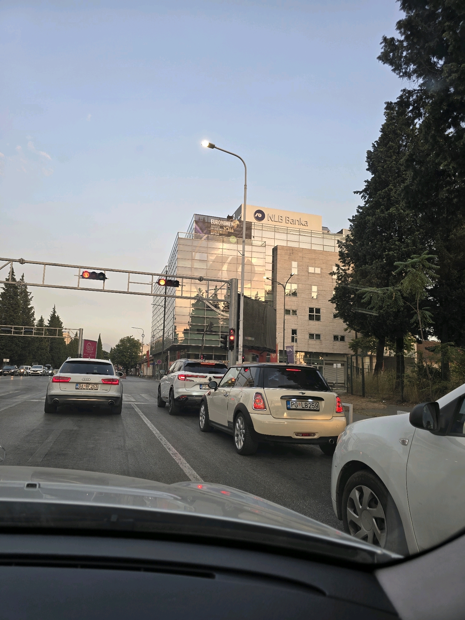 This image captures a street scene in Podgorica, Montenegro, with several cars stopped at a traffic light. In the background, a modern, glass-facade building with the "NLB Banka" logo is visible. The sky is clear with a soft, late afternoon light. Traffic lights are red, indicating a halt in movement. The cars are of various makes and models, with license plates from Montenegro. Trees line the street, and a traffic signal is mounted overhead. The perspective is from inside a vehicle, looking forward.