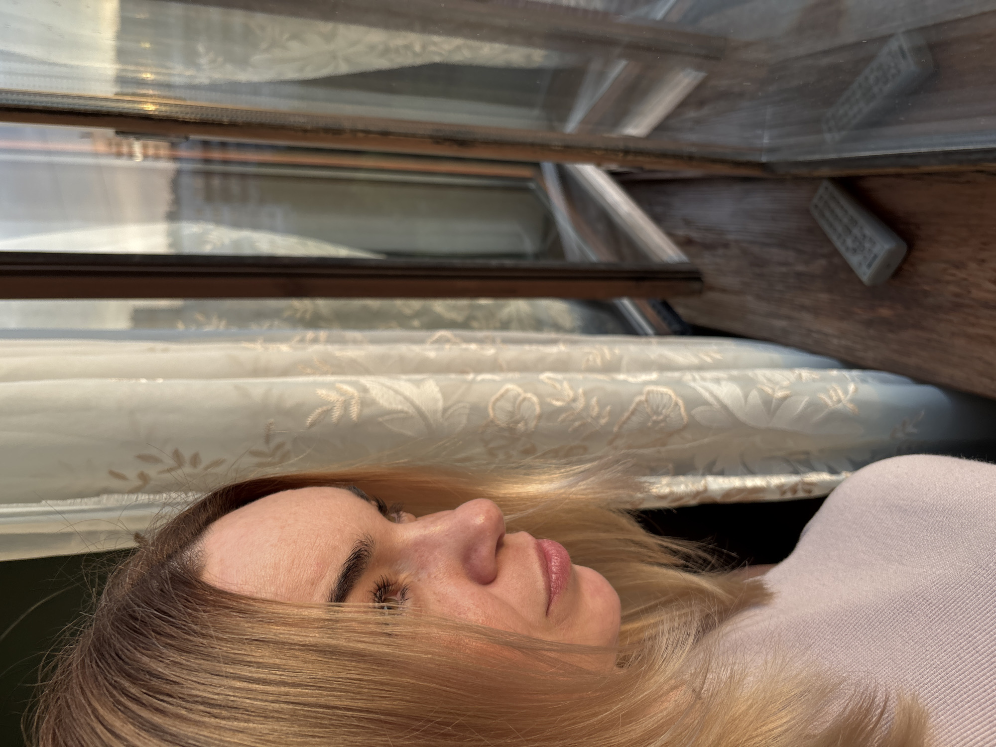 The image displays a woman's profile and surrounding elements, presented in a sideways orientation. In the foreground, on the right side of the frame, a woman is seen from the nose upwards, with her blonde or light brown hair fanned out around her head and partially covering her face. Her right eye is either closed or gazing downwards, and her expression appears calm and serene. Her lips are slightly parted, revealing a soft pink color. She wears a light-colored, possibly knit, garment on her upper body.
Above and to the left of the woman, a window structure is visible, characterized by dark brown wooden or metal frames and multiple glass panes. Behind these frames, a light-colored curtain with an intricate, possibly floral or leafy, pattern can be seen. Sunlight casts warm hues on the woman's hair and skin, suggesting natural daylight, likely from morning or late afternoon. On a wooden surface, possibly a window sill or part of the frame, a small, white or light grey remote control is discernible, though its text is not legible.
The setting is an indoor space, likely a home, with a window providing ambient light. There is no specific activity depicted other than the woman resting or posing in a tranquil manner. No external location cues are visible, and the weather cannot be determined.