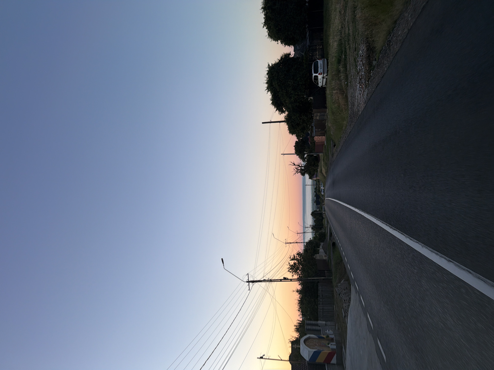 The image depicts a road in the outskirts of Pașcani, Romania, at dawn or dusk. A paved road with a white central line stretches into the distance, curving slightly to the right. Overhead power lines crisscross the sky, supported by utility poles. On the left side of the road, there is a white structure adorned with red, yellow, and blue bands, resembling a monument or memorial, adjacent to a wooden fence. A few houses and trees are visible along the roadside. On the right side, a white car is parked on a grassy area next to a fence, with more trees and houses further back. The sky transitions from a light blue overhead to warmer hues of orange and pink near the horizon, indicating the presence of the sun just below or rising above the landscape. The overall scene conveys a sense of rural tranquility.