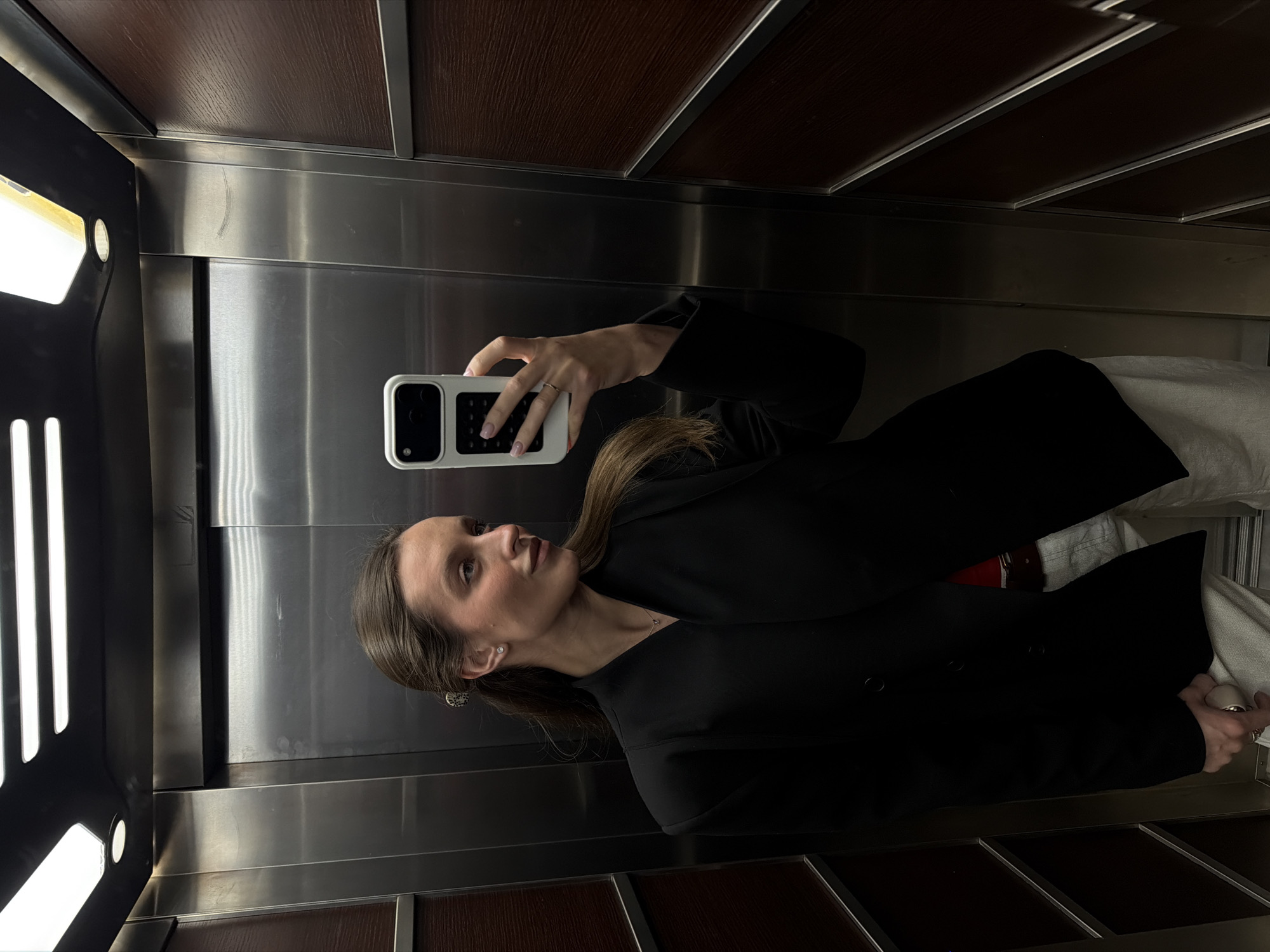 A woman in a black blazer is taking a mirror selfie in what appears to be an elevator with stainless steel walls and brown wooden trim. She is holding a white smartphone with a black and white checkered case. The elevator has vertical lighting strips. The location cannot be determined from the image.