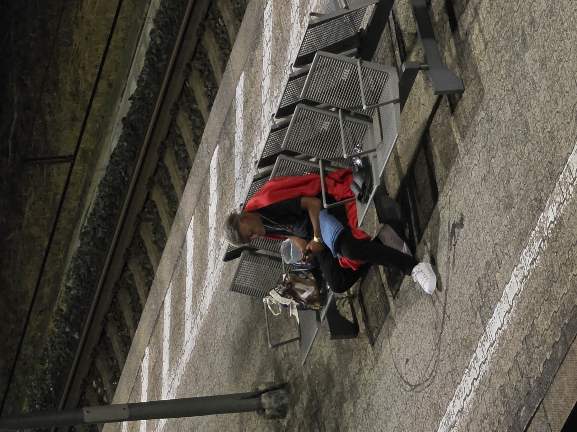 A man sits alone on a metal bench at a train station platform at night. He is wearing a black t-shirt, black pants, and white sneakers. A red cloth is draped over his shoulders. He is looking down at his phone, which he holds in his hands. Next to him on the bench is a bag and a bucket. The train tracks are visible behind him. The platform is paved with stone. White markings on the platform indicate the edge.