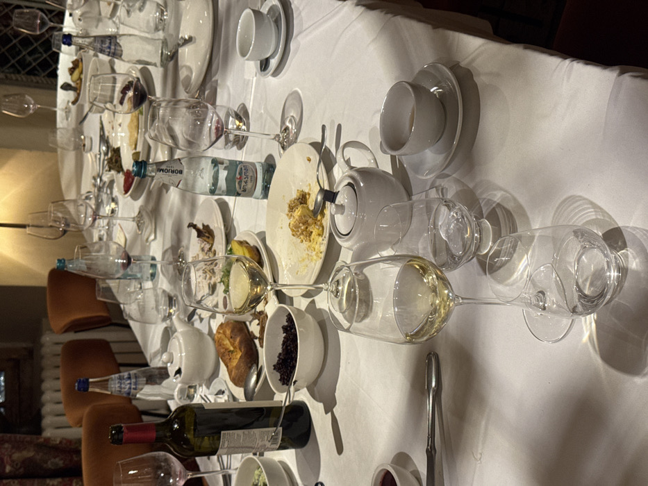 The image depicts a long dining table, covered with a white tablecloth, in an indoor setting, presumably a restaurant or formal dining room, located in Tbilisi, Georgia. The scene captures the aftermath of a meal, with numerous dishes and glassware in various states of use.
Prominently displayed are multiple wine glasses, some containing residues of red wine, others with white wine. Several water glasses, some partially filled, are also present. A mostly empty dark green bottle of red wine stands on the left. Bottles of mineral water, specifically "Borjomi," a Georgian brand, are visible.
The table features numerous white ceramic plates, many containing remnants of food, indicating a recently concluded meal. Visible food items include a partially consumed piece of savory bread or pastry, a bowl filled with dark, small food items (possibly berries or grains), and other plates with yellowish and greenish food residue. Several white ceramic teapots and teacups with saucers are also distributed across the table. Serving spoons and forks are scattered among the dishes.
The background is dimly lit, revealing upholstered chairs with orange-brown backs. A white wall radiator is visible on the left. A lamp provides warm illumination in the distant background, and a window with a visible frame or grille pattern can be discerned. No individuals are present in the immediate foreground.