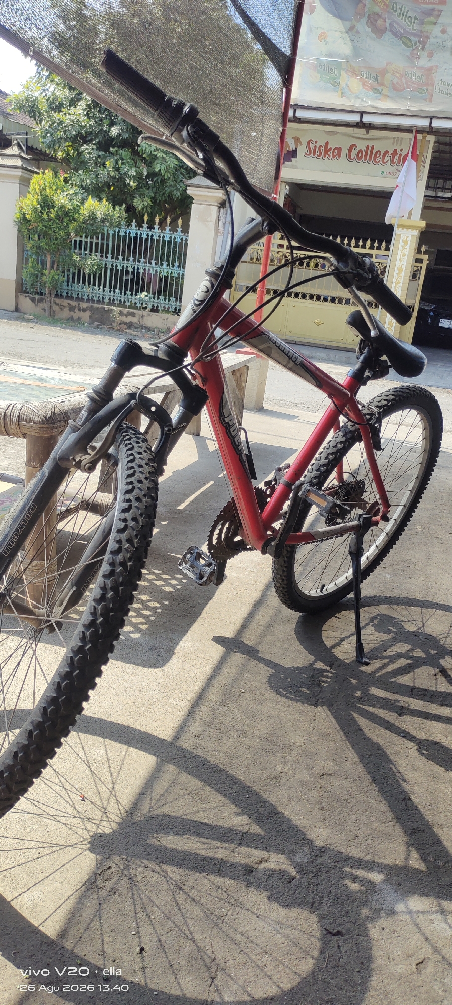 A red and black bicycle is parked on a concrete surface, casting a long shadow. The bicycle is the main subject, with its front wheel and handlebars visible in the foreground, and the rest of the frame extending towards the background. The background reveals a building with a sign that reads "Siska Collection" and displays advertisements. A white and red Indonesian flag is also visible. Behind the building, a green metal fence and some trees are present. The overall scene suggests an outdoor setting, possibly a street or a shopfront in Simo, Indonesia. There are no people visible in the image.