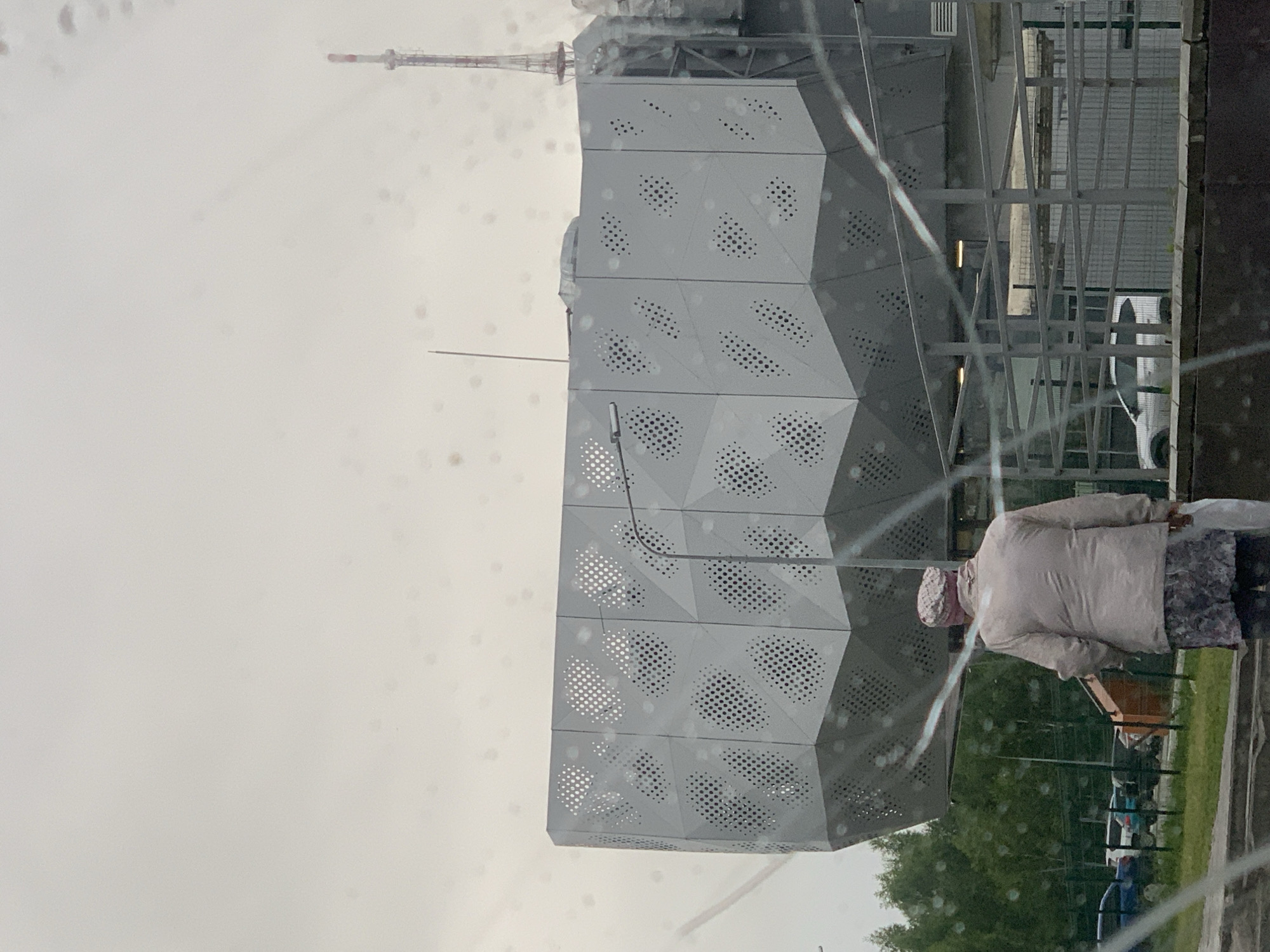 The image shows a person from behind, standing near a modern building with a geometric, perforated facade. The building's exterior is composed of angled panels with a pattern of circular holes. To the left of the building, a radio tower or similar tall structure is visible against an overcast sky. The person is wearing a light-colored jacket and a patterned head covering, and a patterned skirt or trousers. They are holding a white bag or object in their right hand. In the background, there is a grassy area with trees and a fence. The overall scene suggests an outdoor setting. The presence of water droplets on the surface from which the photo is taken indicates that it is raining or has recently rained. The lighting is diffused, consistent with an overcast day. The specific location of Nizhnekamsk, Russia, provides geographical context.