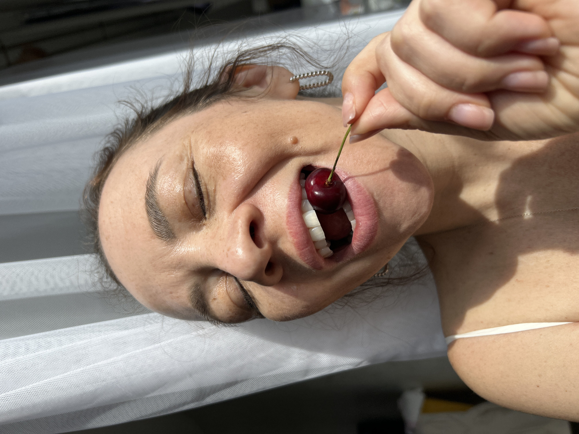 A woman is positioned on a white, translucent surface, possibly a hammock or bed. She holds a dark red cherry by its stem, placing it in her mouth. The woman's eyes are closed, and her face is tilted towards the sun. She wears a thin silver earring. A thin silver necklace is visible on her chest. The background is out of focus. There is no explicit evidence to support the location being in Chaikovsky, Russia.