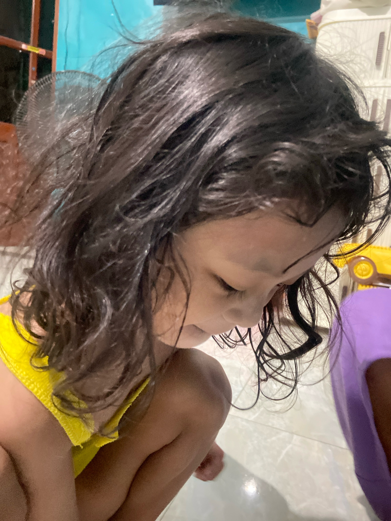 This close-up image shows a young child with dark, wet hair, seen from the side and slightly from above. The child is wearing a yellow top and is in a crouched or kneeling position. Their head is bowed, and their eyes are closed or looking downwards, suggesting they are either concentrating, resting, or possibly upset. The wet hair is spread around their face and shoulders, with some strands falling forward.
In the background, there are hints of a domestic setting. To the left, a fan with a circular grille is visible, and behind it, a turquoise wall and a wooden frame suggest an interior space. To the right, a portion of a white cabinet or storage unit is visible, along with a bright yellow toy with circular wheels, and a large purple object. The floor appears to be tiled. The overall lighting suggests it is daytime indoors. The child’s posture and the wet hair might indicate they have recently bathed or played with water. The scene is likely in Gelam, Indonesia.